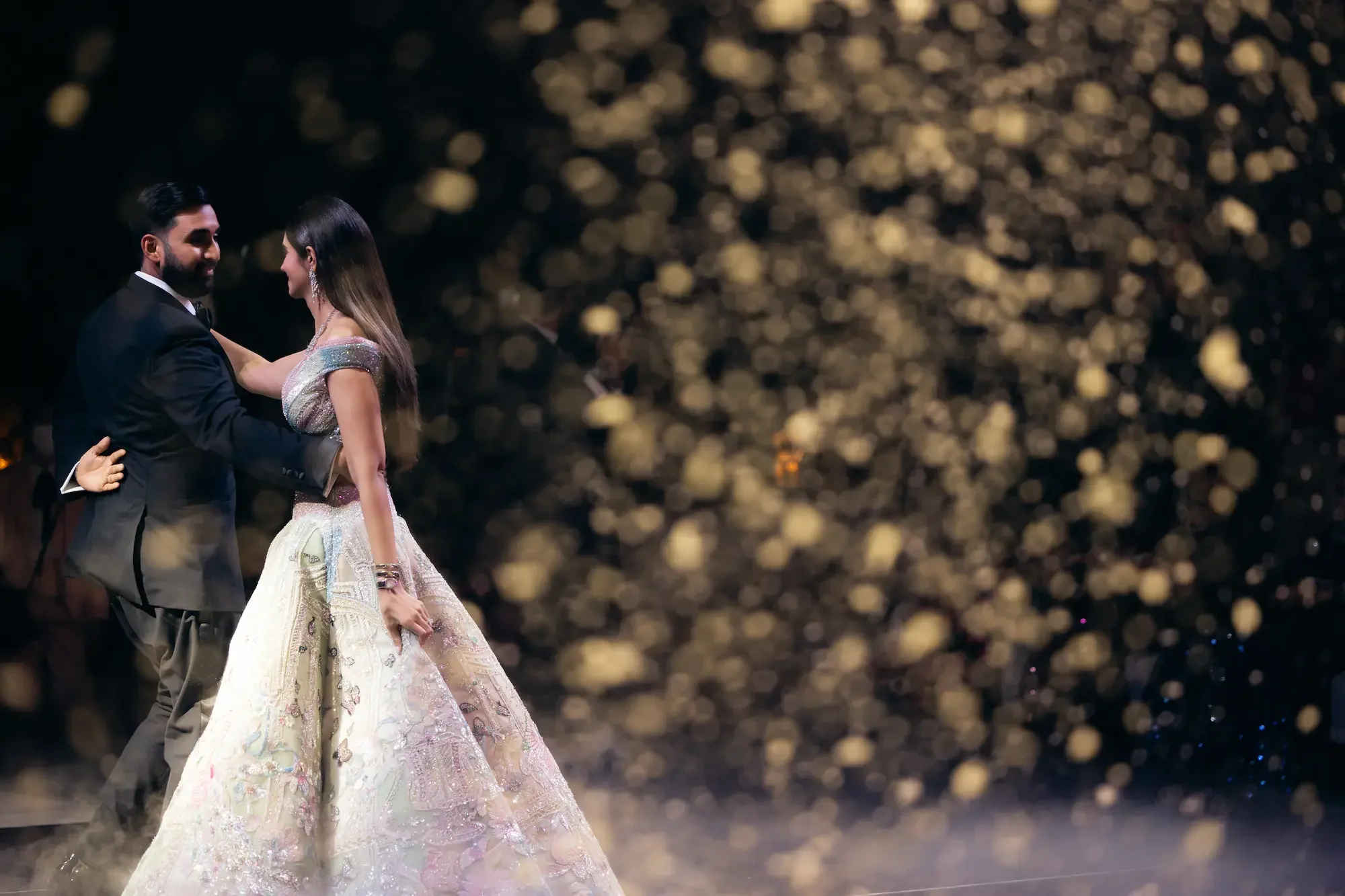 A couple in formal attire dancing at a wedding reception, with golden bokeh lights in the background.