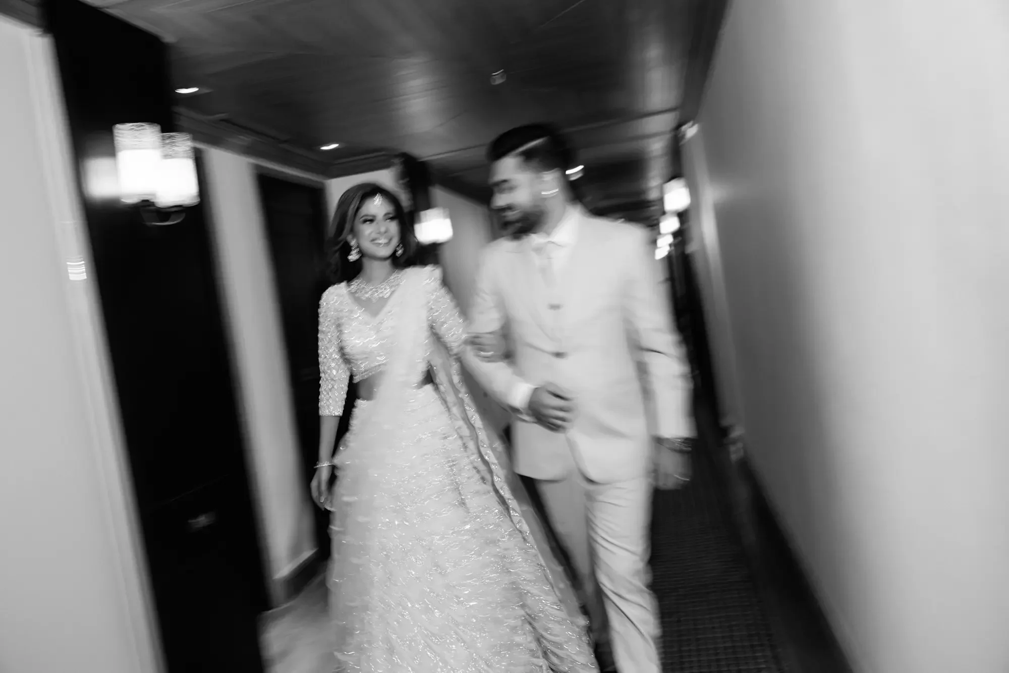 A woman in a sparkling gown and a man in a suit walking down a hallway, smiling and holding hands.