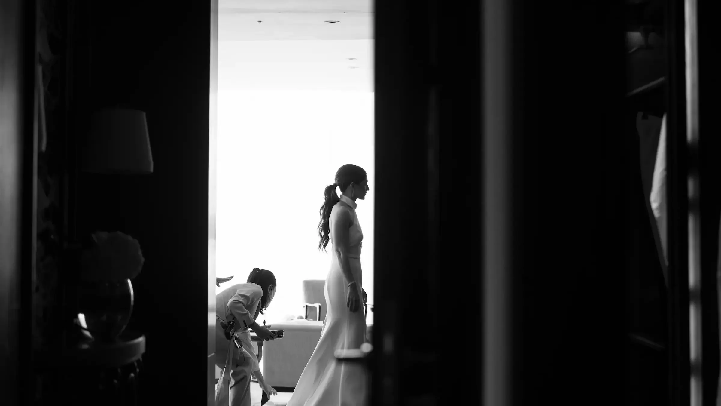 Black and white photo of a woman in a dress walking in a room with two children. The image is viewed through an opening, with objects partially obstructing the view.