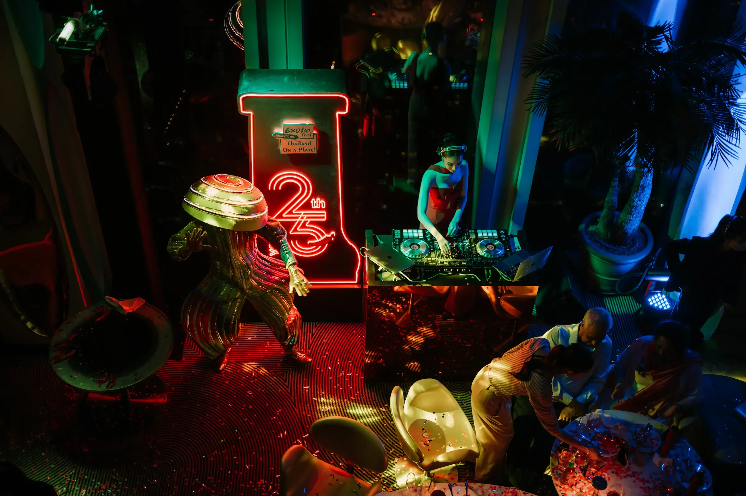 A nightclub scene with a DJ wearing headphones at a mixing console, colorful neon lights including a large neon sign with the number 25 and the words "Exotic Food, Thailand, On a Plate!", a chrome alien statue, and tropical plants, with some people dancing and socializing.