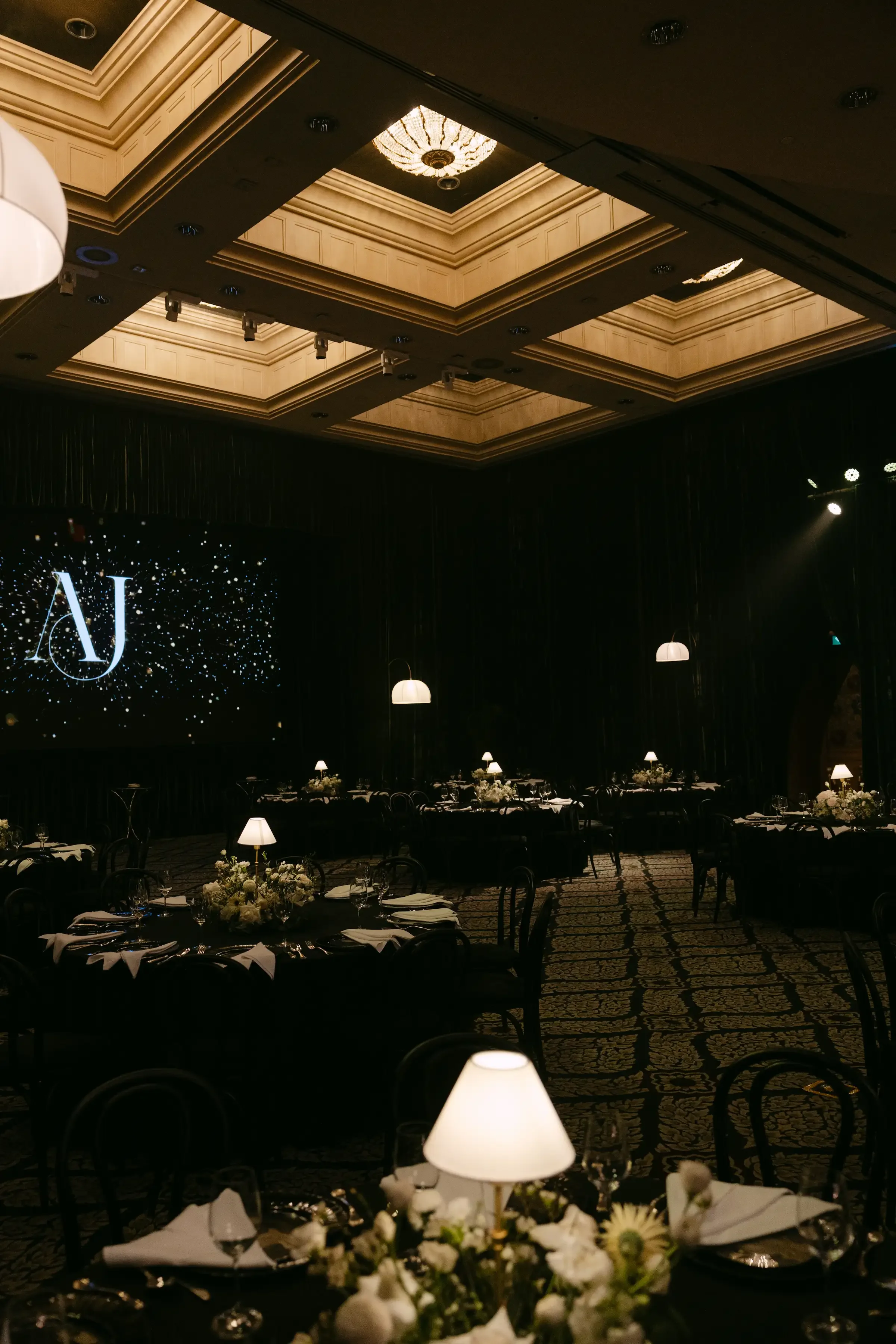 An elegantly decorated banquet hall set for a formal event with round tables, white floral centerpieces, black tablecloths, and glassware. The room features a decorated ceiling with a chandelier, dark curtains, and dim lighting. There is a large screen displaying a logo with the initials 'NJ' and a dark background.
