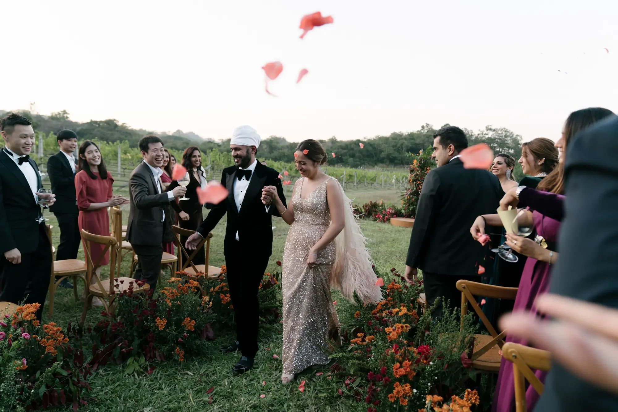 A wedding celebration outdoors with a bride and groom holding hands, surrounded by friends, with flower petals falling from above.