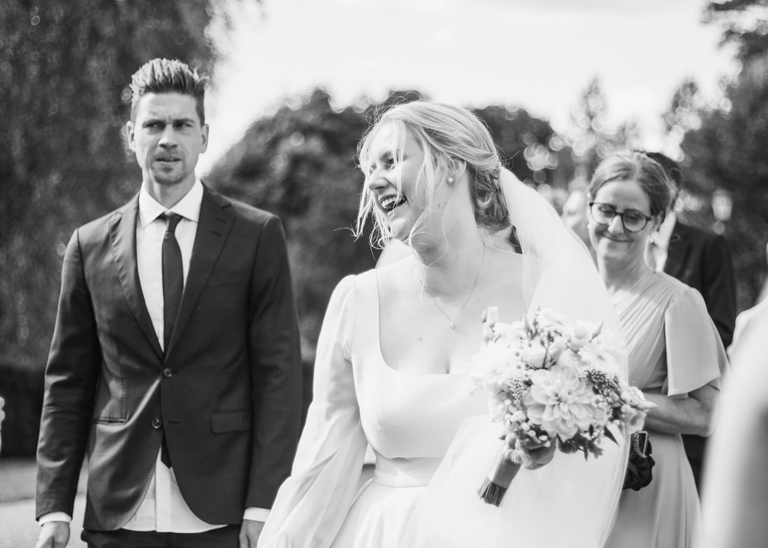 Bröllopsfoto i svartvitt föreställande en glad brud med blont hår och vitt bröllopsklänning som håller en bukett, en man i kostym och slips, och en kvinna med glasögon och mörkt hår i bakgrunden, utomhus.