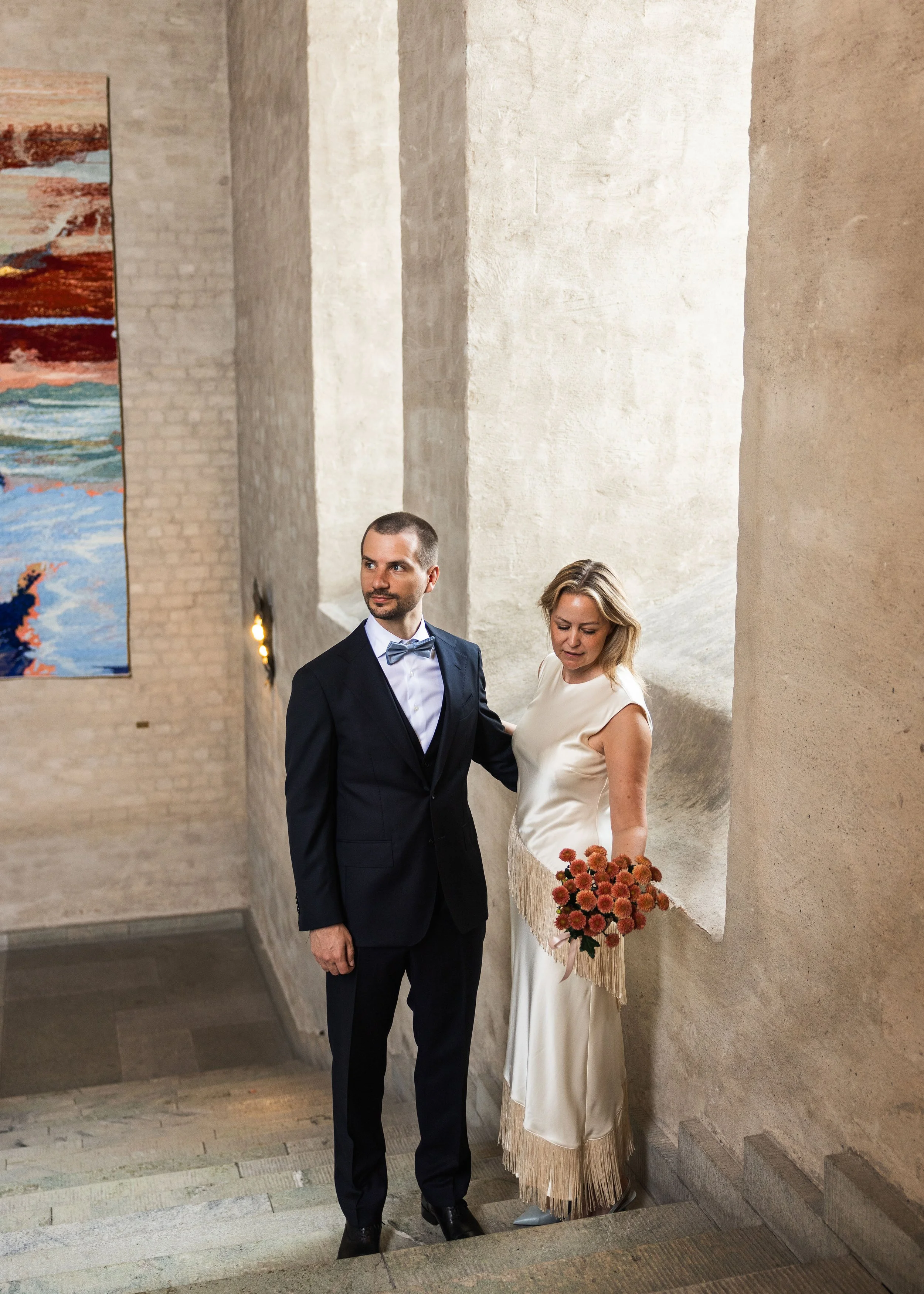 En man i mörk kostym och en kvinna i vit brudklänning står tillsammans i en ljus, modern hall med konst på väggarna och trappsteg av sten.