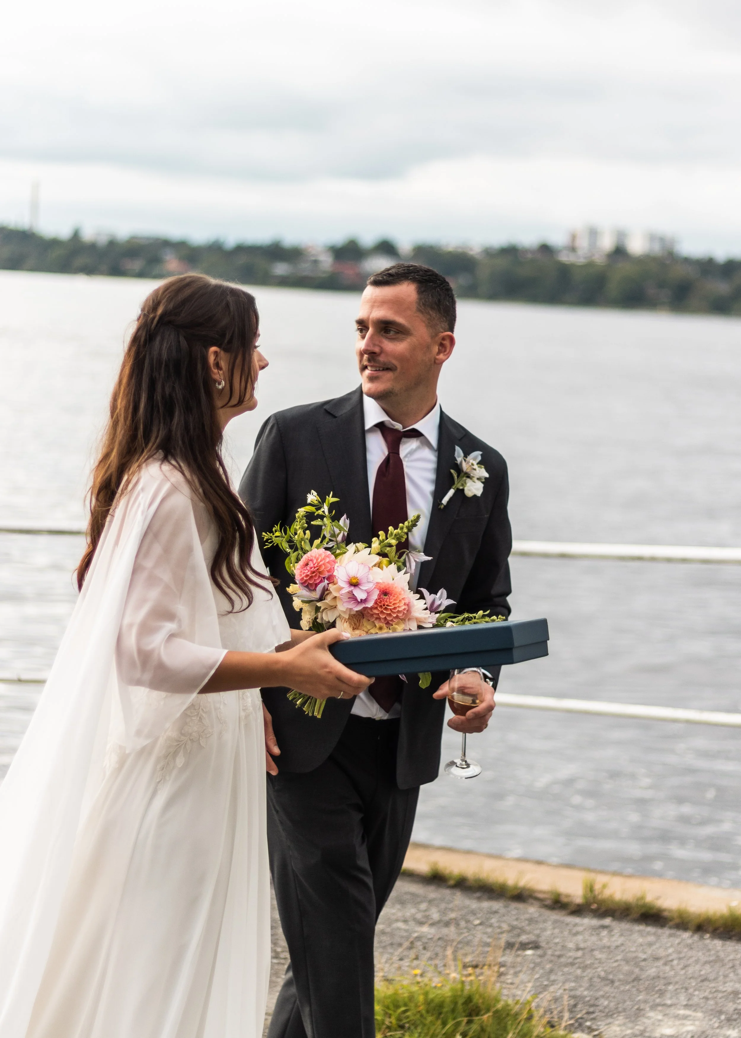 En brud och en brudgum står vid vattnet, brudgumen ger bruden en bukett blommor, han håller även ett glas vin i handen.