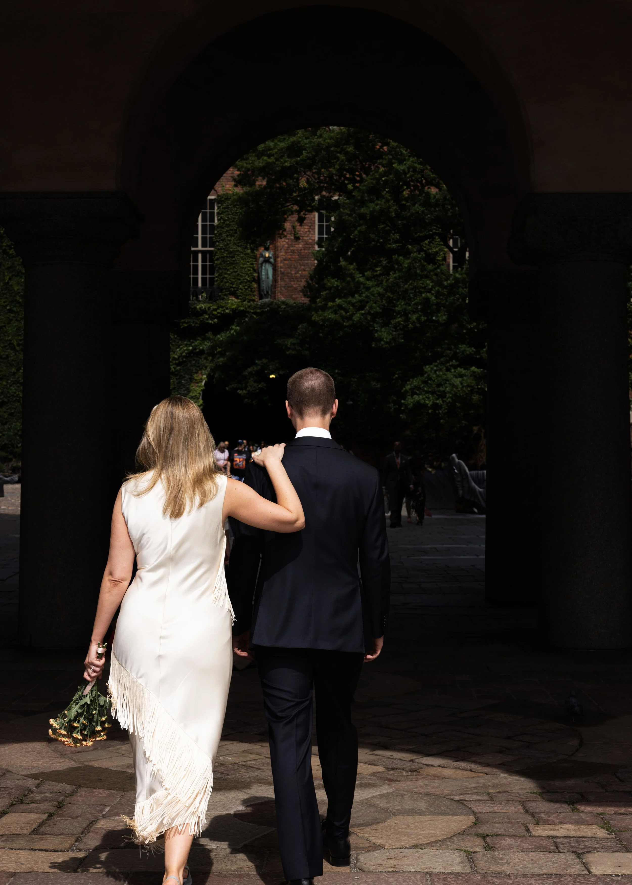 En man och en kvinna går hand i hand under en stenbåge, kvinnor håller en blomsterkvast i handen, andra människor i bakgrunden, grönt träd och en byggnad syns i bakgrunden.