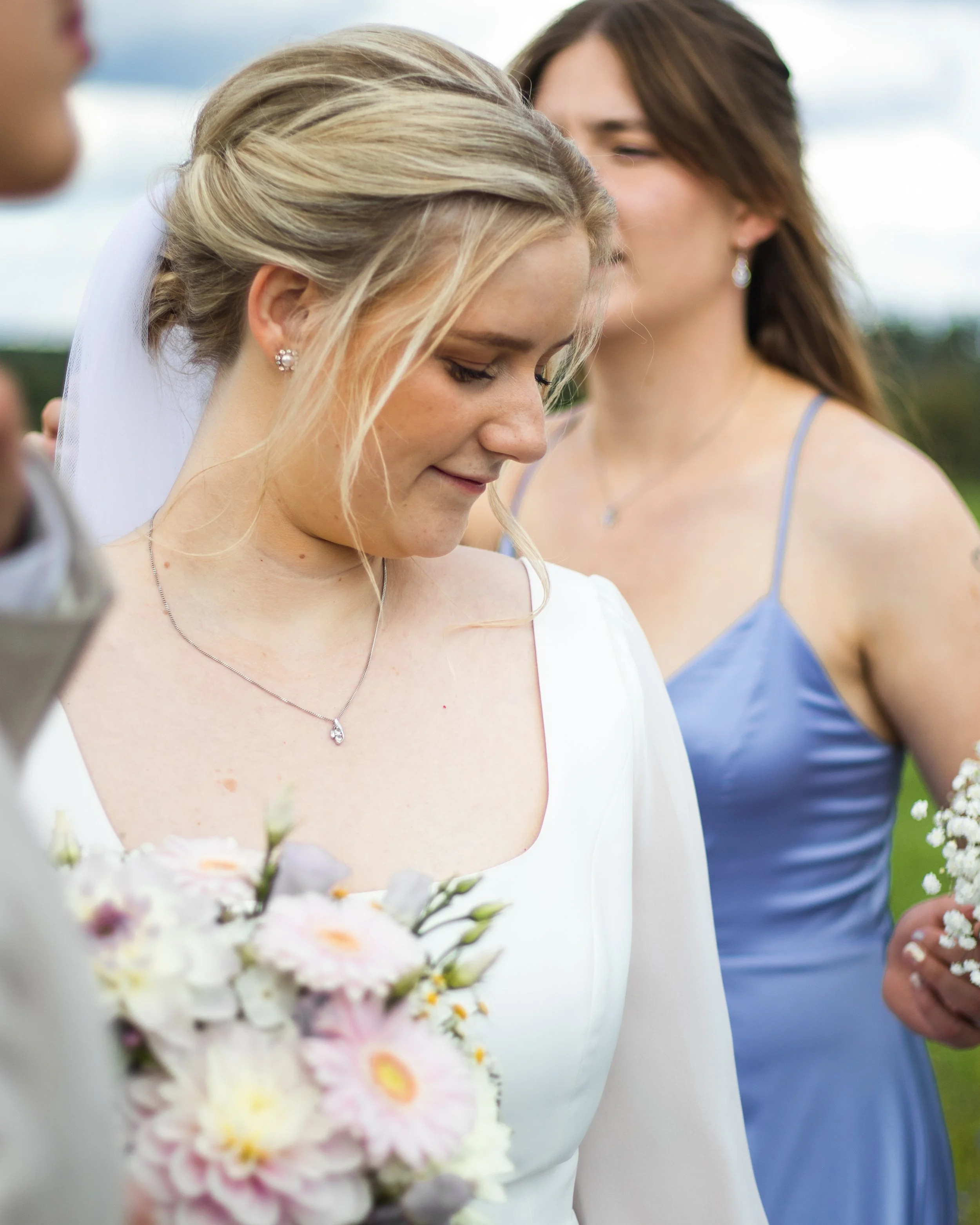 En brud med blonda lockigt hår i brudklänning och ett par pärlörhängen. Hon har ett silverhalsband med ett droppformat smycke. En vän eller tärna i blått klädesplagg står bakom henne. Både finns ett blomsterarrangemang i förgrunden.