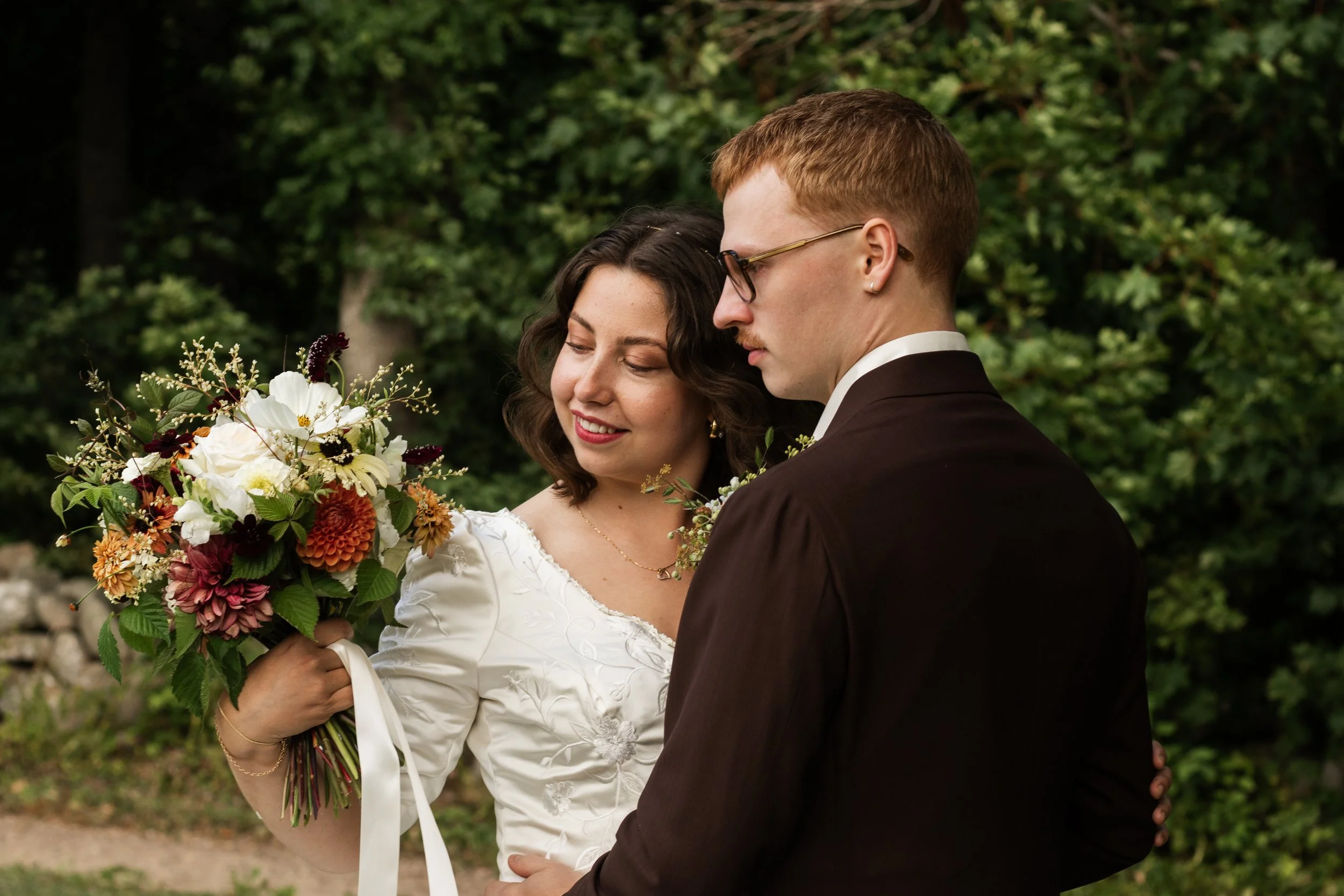En brud och brudgummi som står nära varandra utomhus, omgivna av gröna träd, eventuellt under en ceremonin. Bruden är klädd i vit klänning och håller en stor bukett blommor, medan brudgummen har på sig en mörkbrun kostym med glasögon.