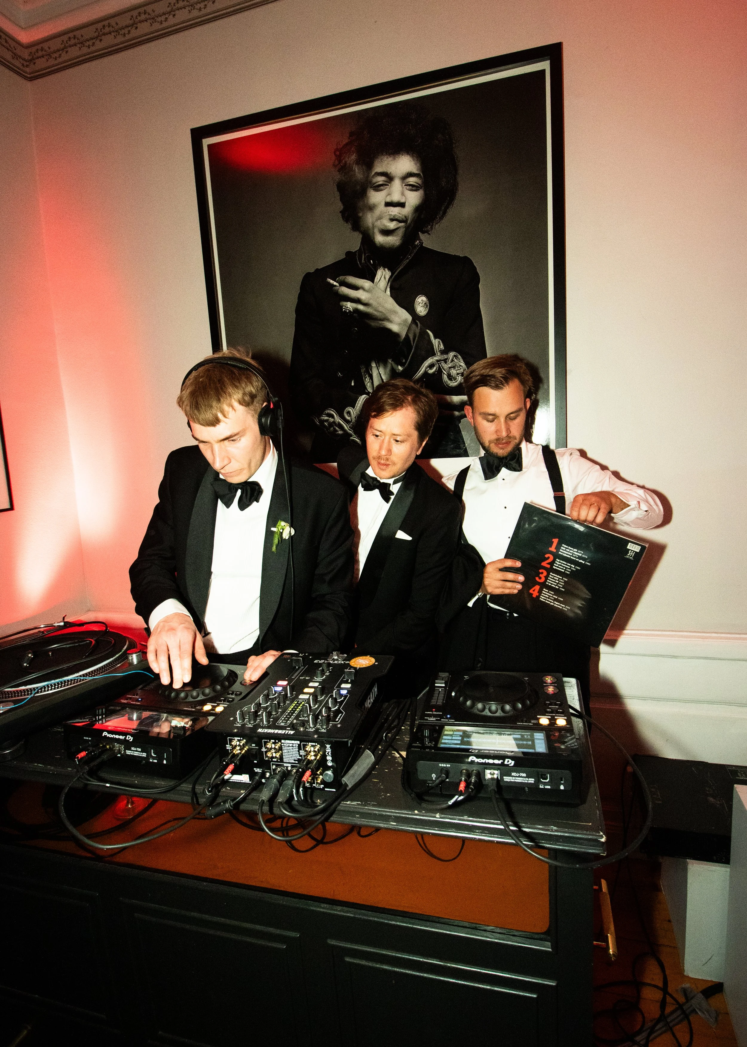Tre män i svart smoking, en med hörlurar, en med en meny, står bakom DJ-utrustning vid ett event. Stort svartvitt porträtt av en man med afrofrisyr och cigarett hänger på väggen.