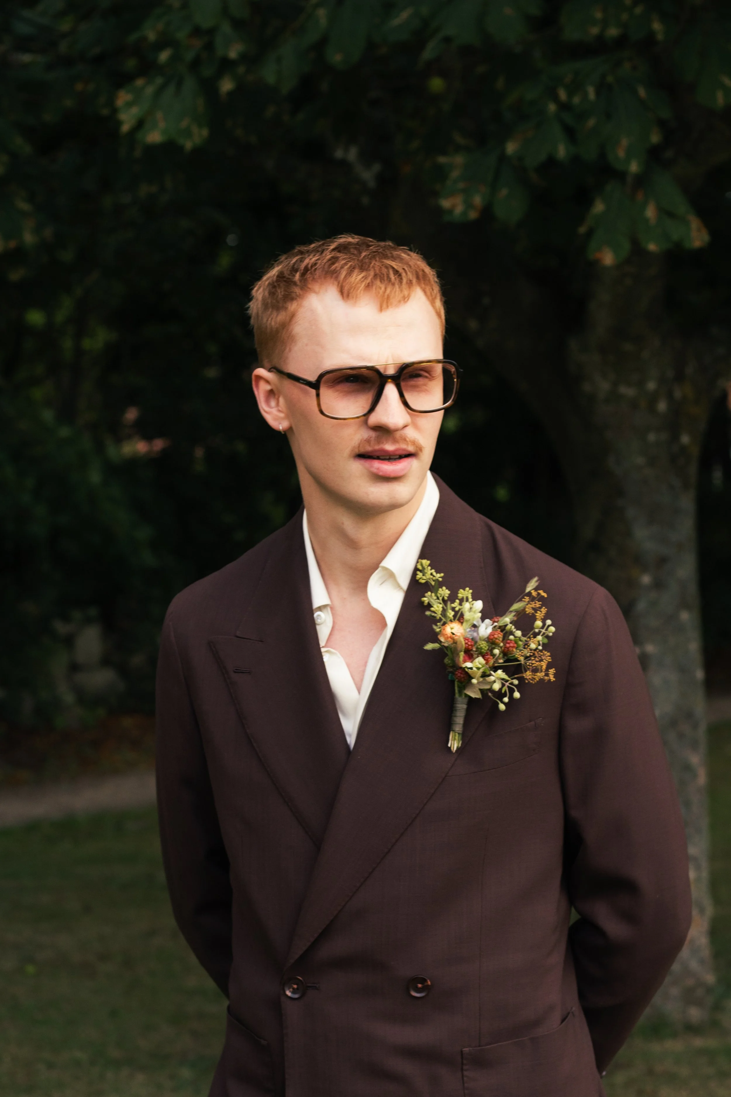 En ung man med rött hår, glasögon, mörk kostym och välsmakande blomsterhalsband står utomhus i en park eller trädgård.
