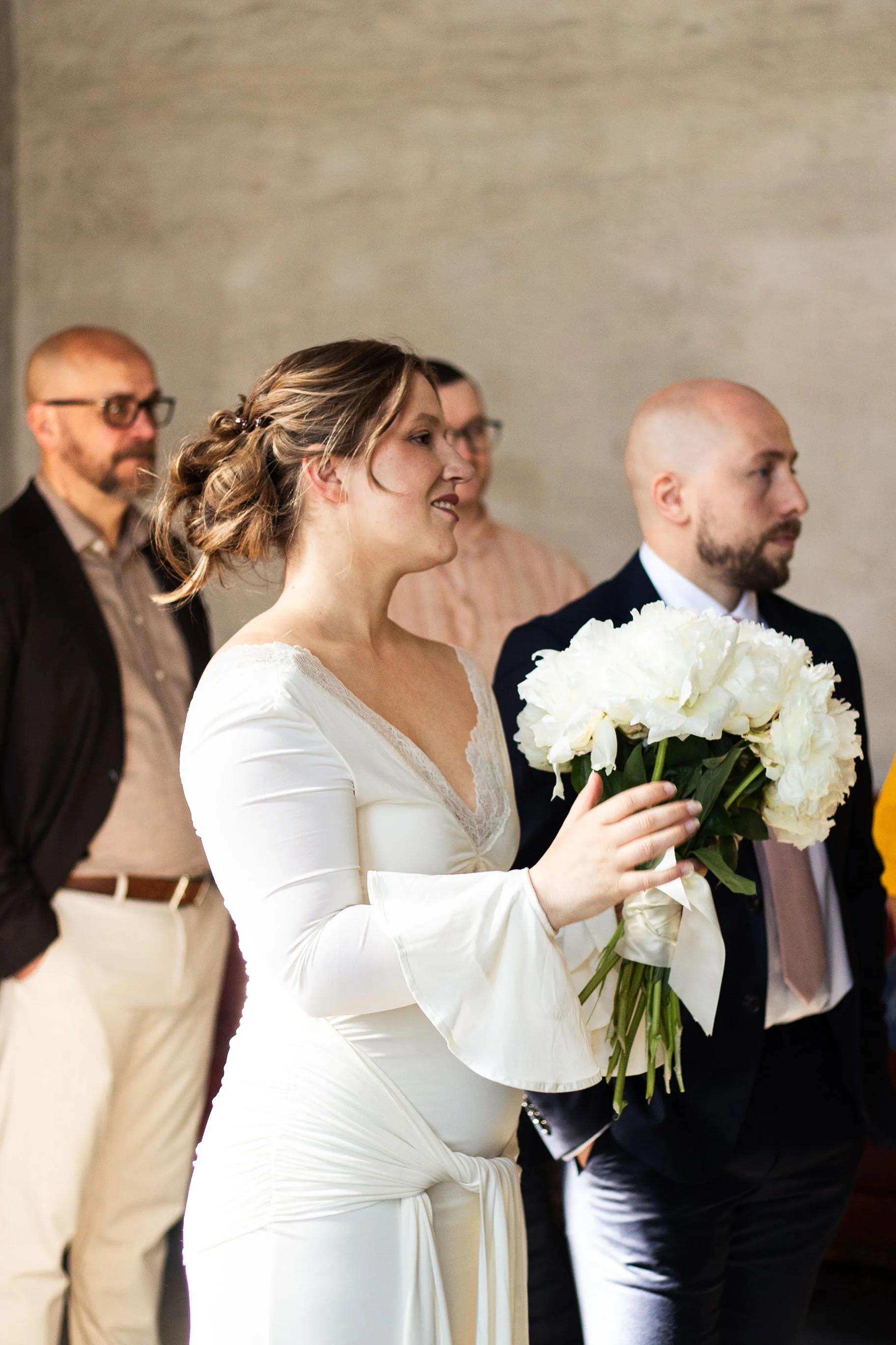 En brudklädd kvinna i vitt håller en stor bukett vita blommor under ett bröllopsceremoni, omgiven av gäster.