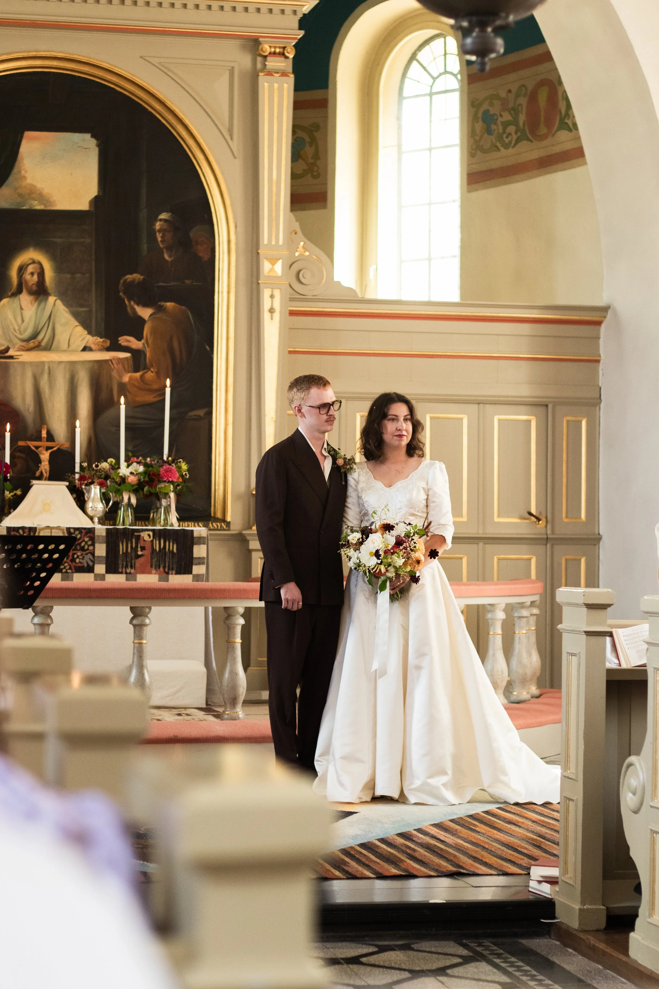 En brud och en brudgum står tillsammans i en kyrka under ett bröllop, med en religiös bild av Jesus i bakgrunden och ljus som strömmar in genom fönstret.