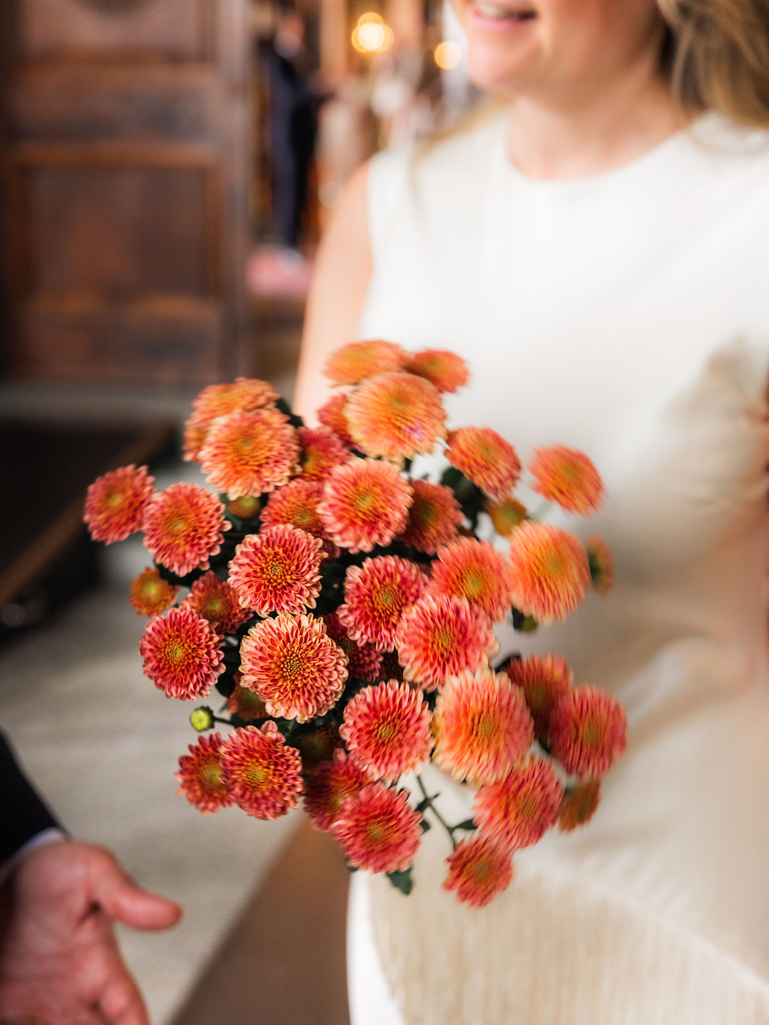 En person håller i en bukett orangea och rosa blommor, personen är klädd i vit klänning och är i ett inomhusmiljö.