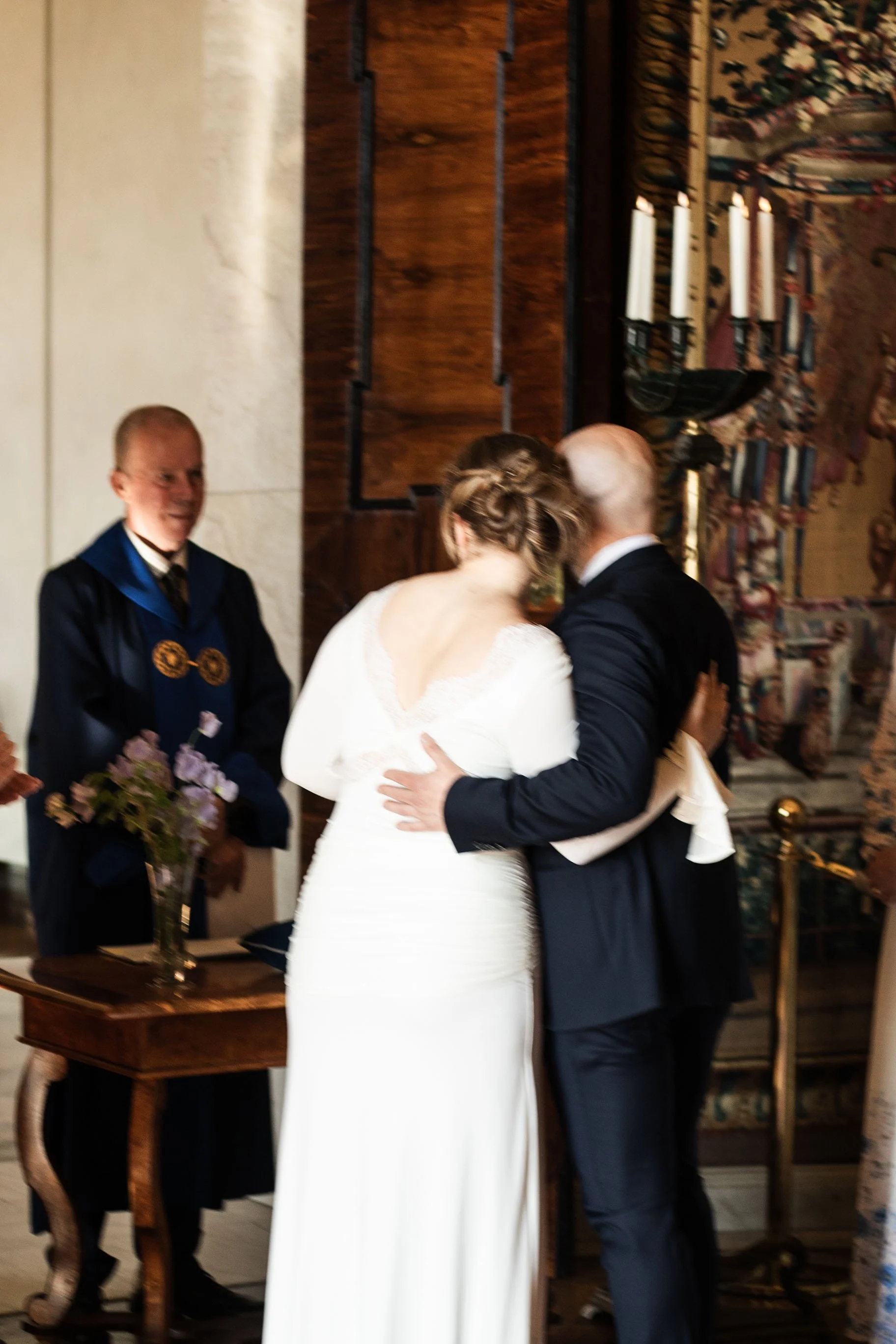 En brud och en brudgum kysser varandra under ett bröllopsceremoni i en kyrka, med en ceremonivärd i bakgrunden och ett altare med ljus i bakgrunden.