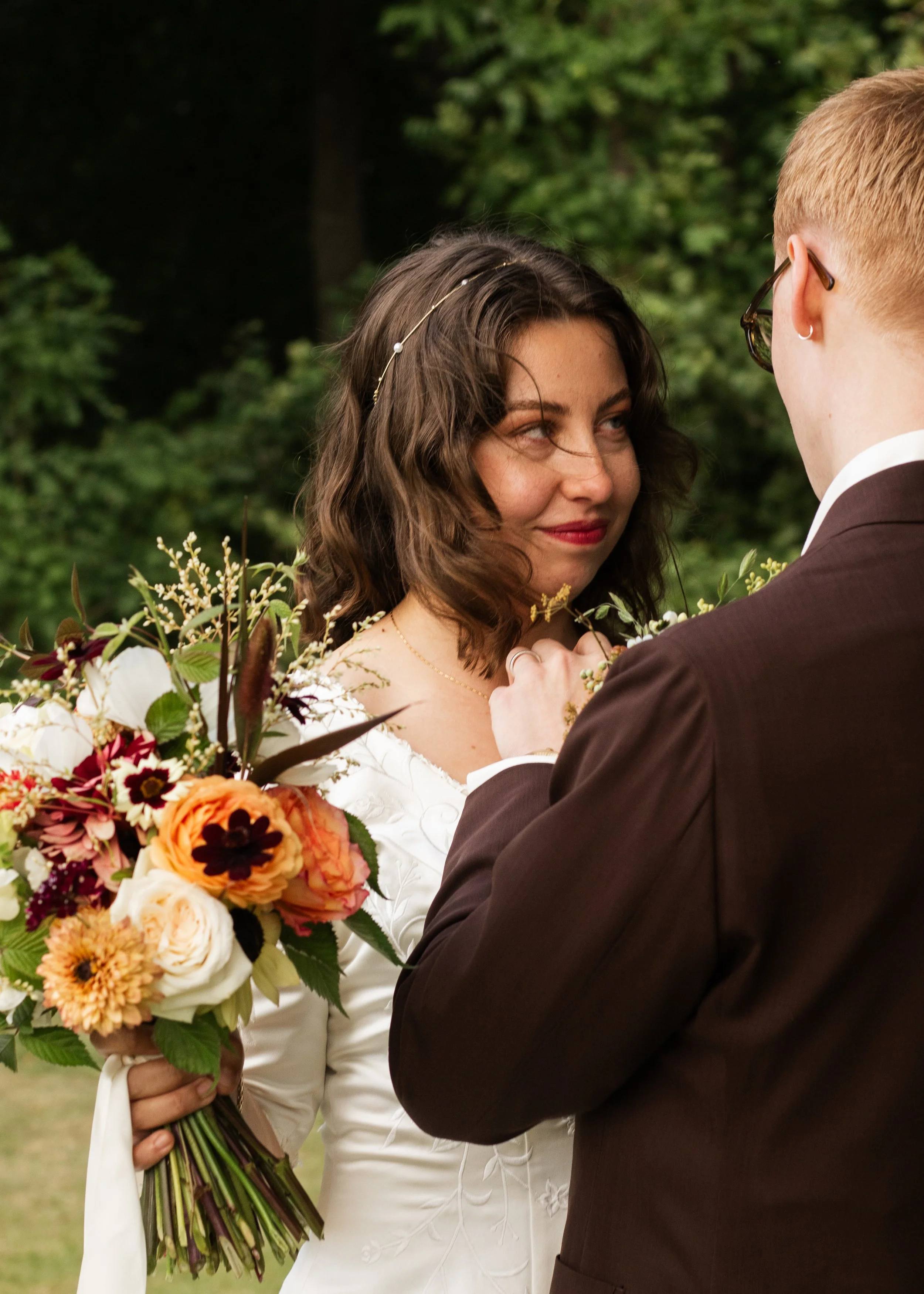 En brud och en brudgum som är ute på deras vigsel, med brudens bouquet av blommor i handen, i en utomhusmiljö med gröna träd i bakgrunden.