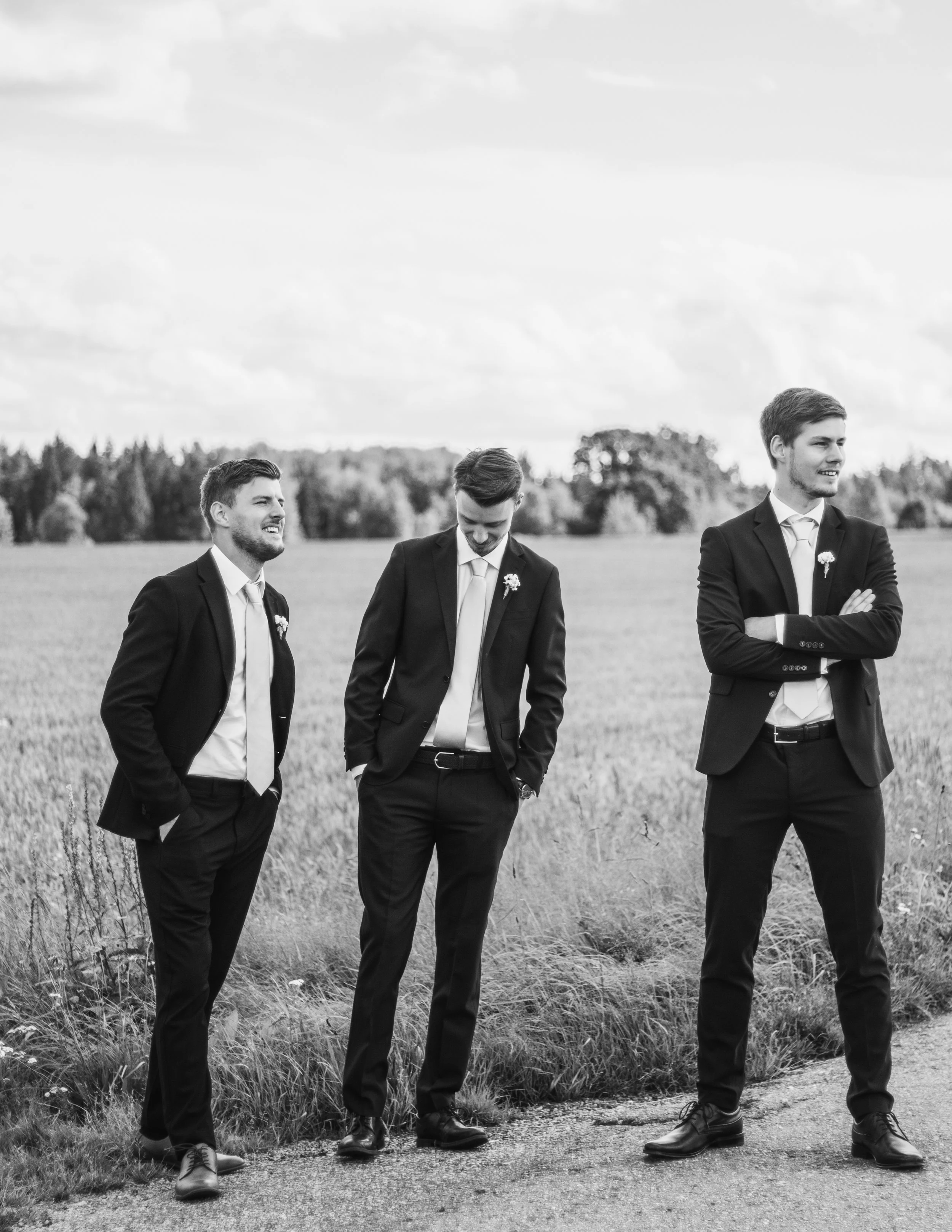 Tre män i mörka kostymer står i en grusväg med fält och träd i bakgrunden, alla har vita skjortor och slips, och två av dem har boutonnéer. De ser glada ut.