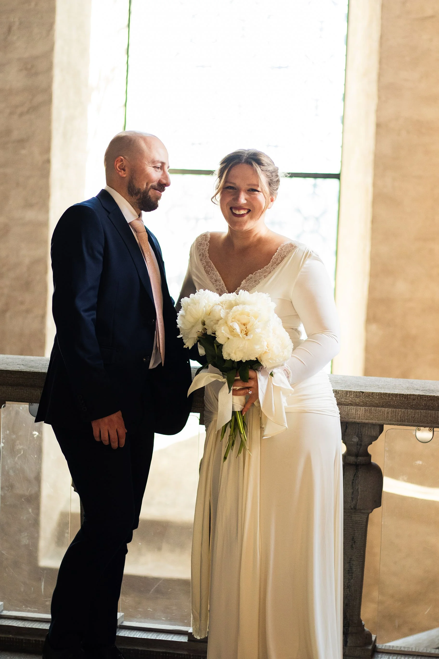 en man i mörk kostym och en kvinna i bröllopsklänning som håller en bukett med vita blommor, står inomhus framför ett fönster.