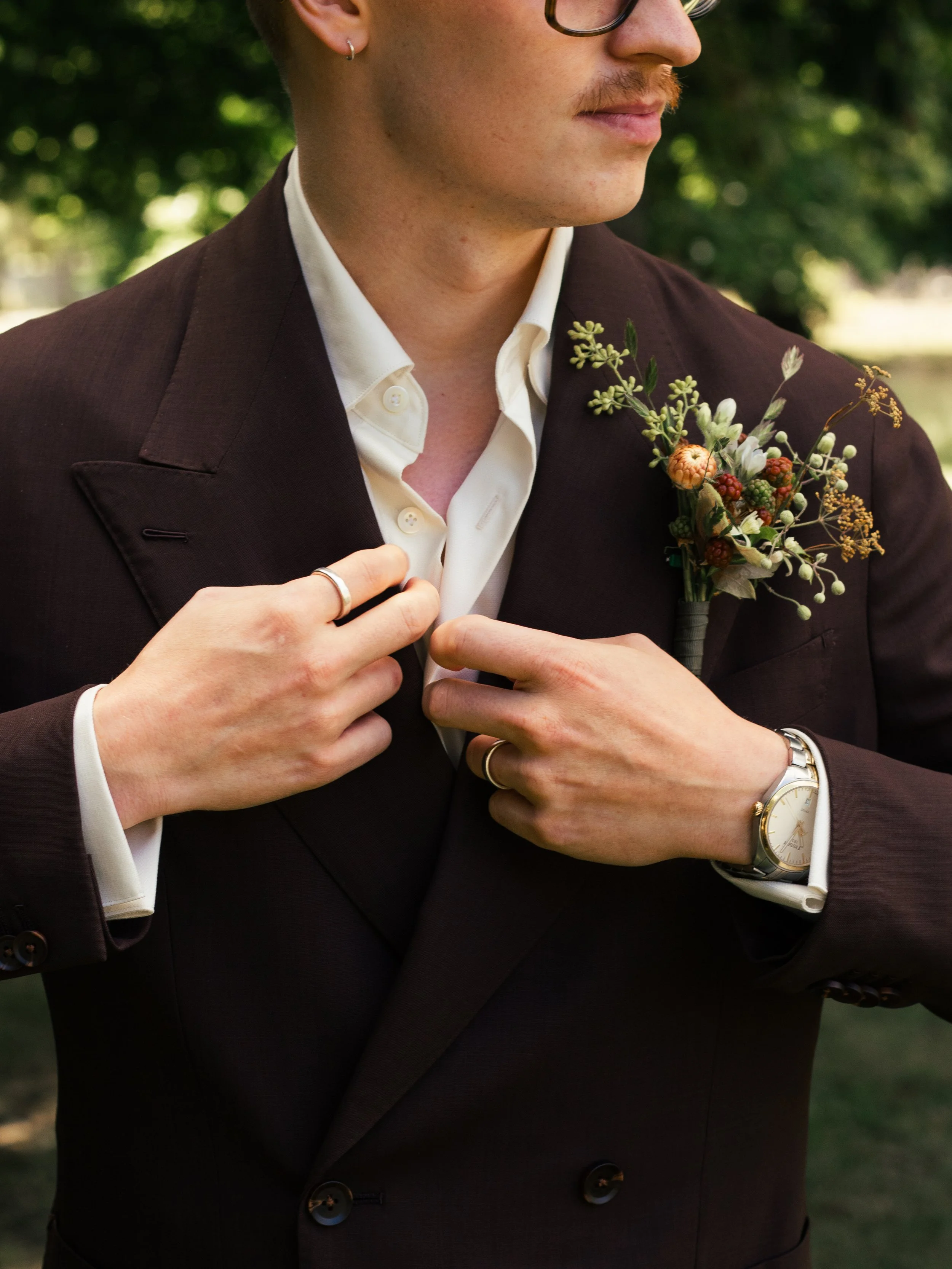 En person i mörk kostym med vita skjorta, bär en klocka och ett ring, och har en blomsterboutonnière på kavajslaget, utomhus med grön bakgrund.