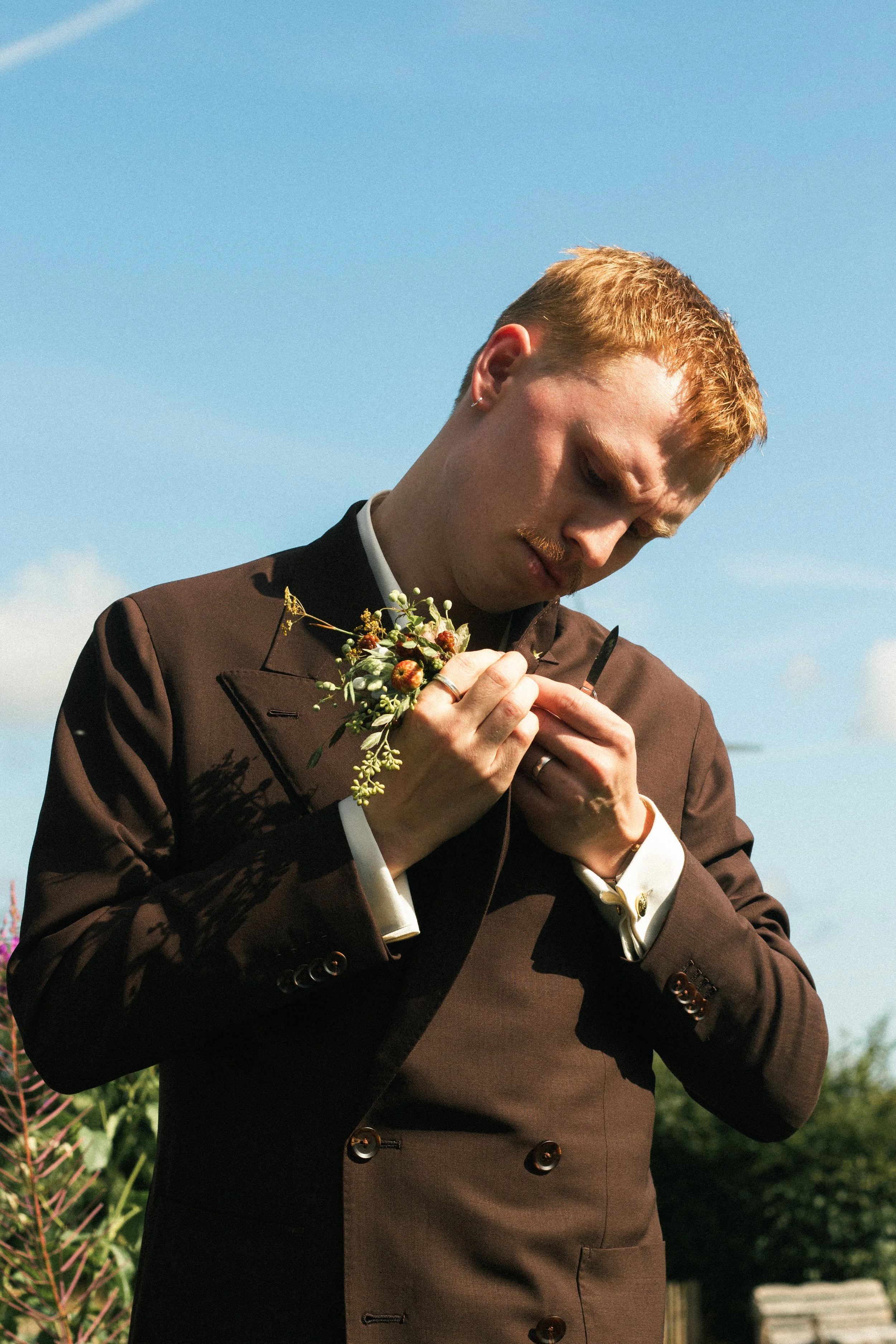 En person med rött hår och mustasch, klädd i en brun kavaj, håller en liten bukett med blommor och klämmer en kniv, står utomhus mot en blå himmel.