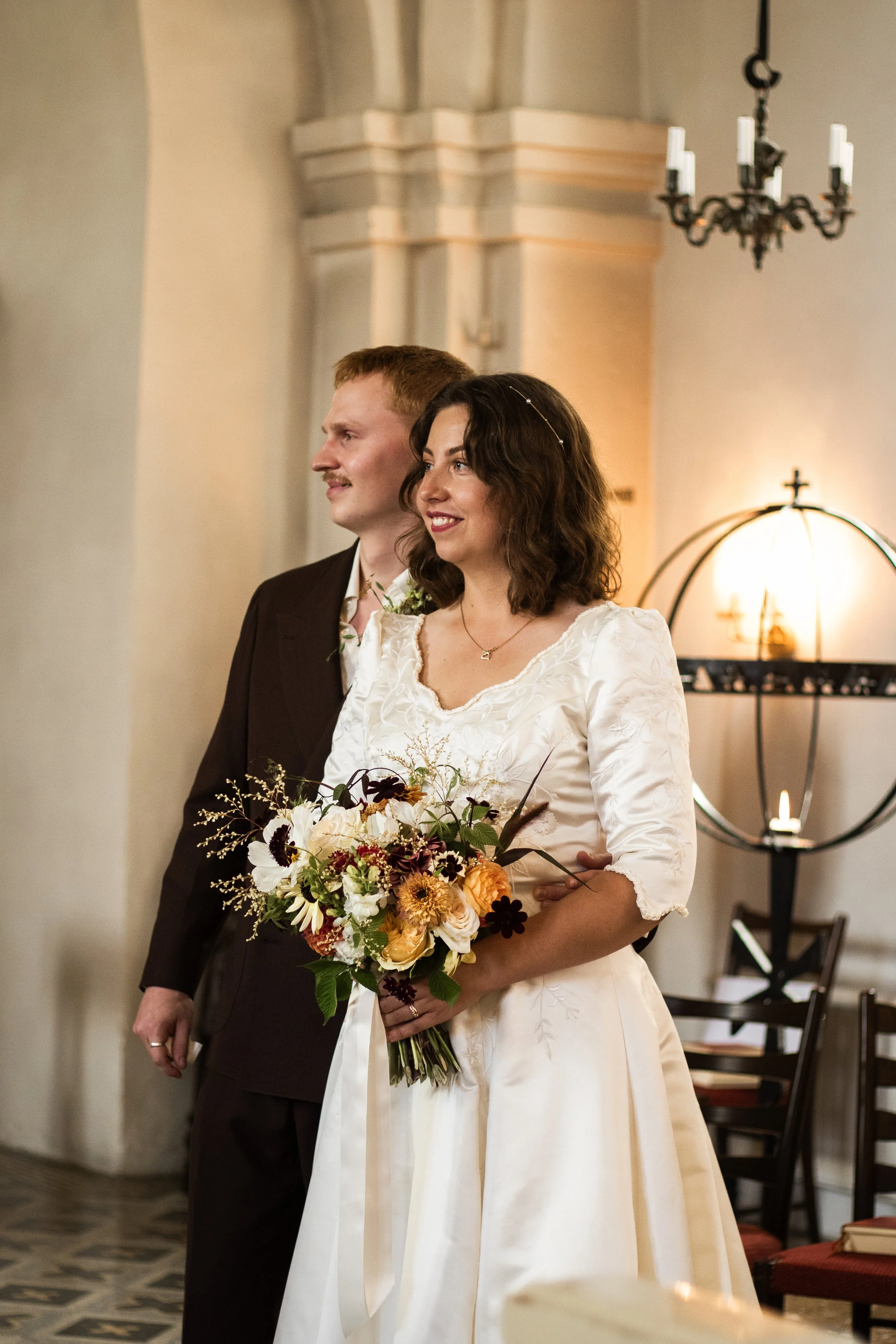 En brud och en man står bredvid varandra under ett bröllopsceremoni i en kyrka. Bruden bär en vit brudklänning och håller en stor bukett med blommor, medan mannen är klädd i en mörk kostym.