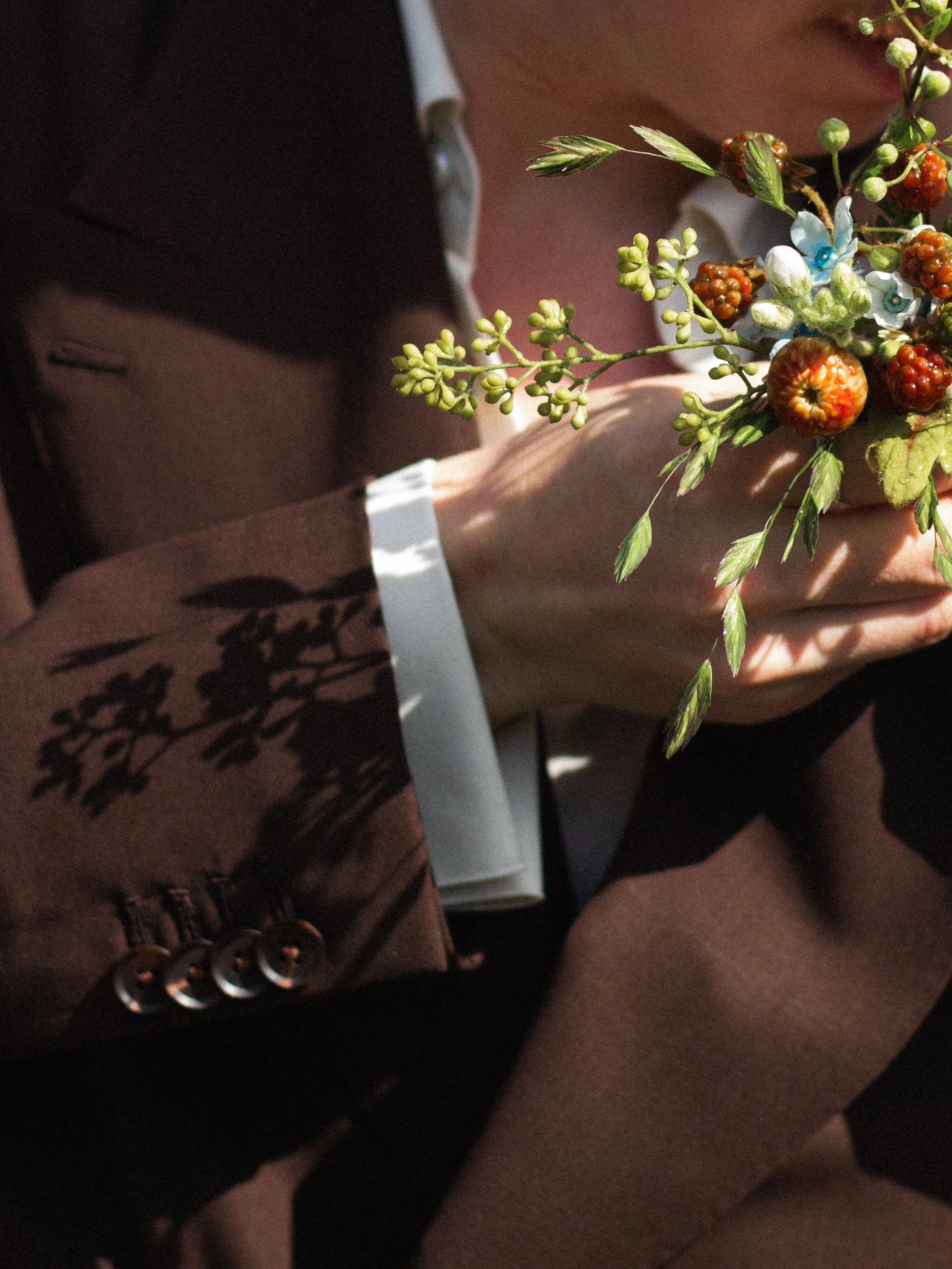 En brudgum håller i ett blomsterarrangemang med små gröna bär, grönt blad och blå blommor, klädd i svart och mönstrad kjol.