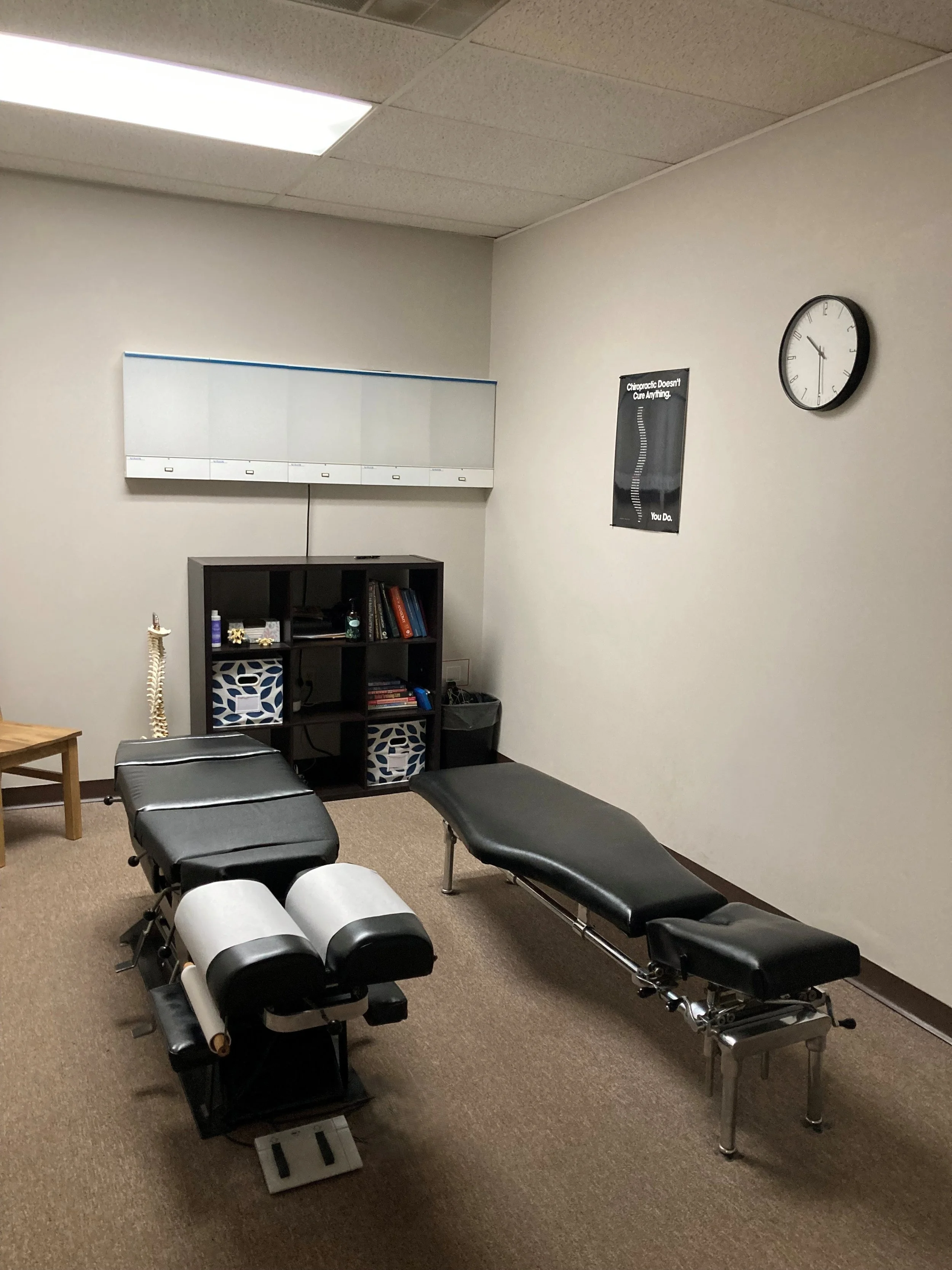 A chiropractor's office with a chiropractic adjusting table, a shelf with books, posters, a clock, and exercise equipment.