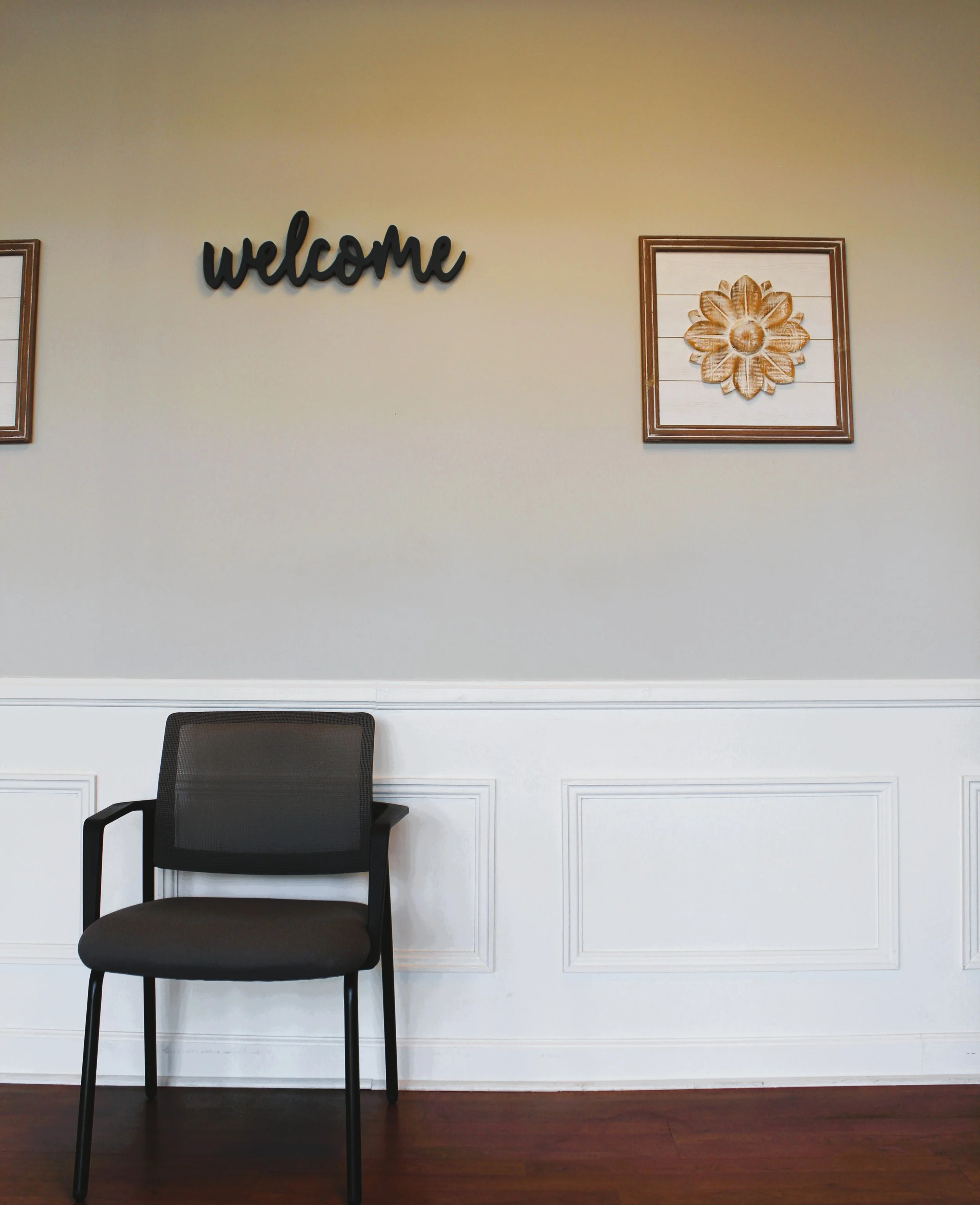 An empty black office chair with armrests in front of a white wainscoted wall. The wall has decorative framed art, including a framed flower, and a black cursive 'welcome' sign.
