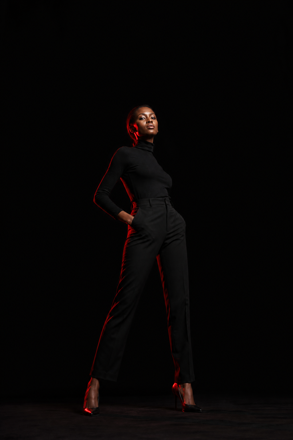 A woman in a black turtleneck, black high-waisted pants, and black heels standing with her hands in her pockets against a black background, with subtle red lighting highlighting her silhouette.