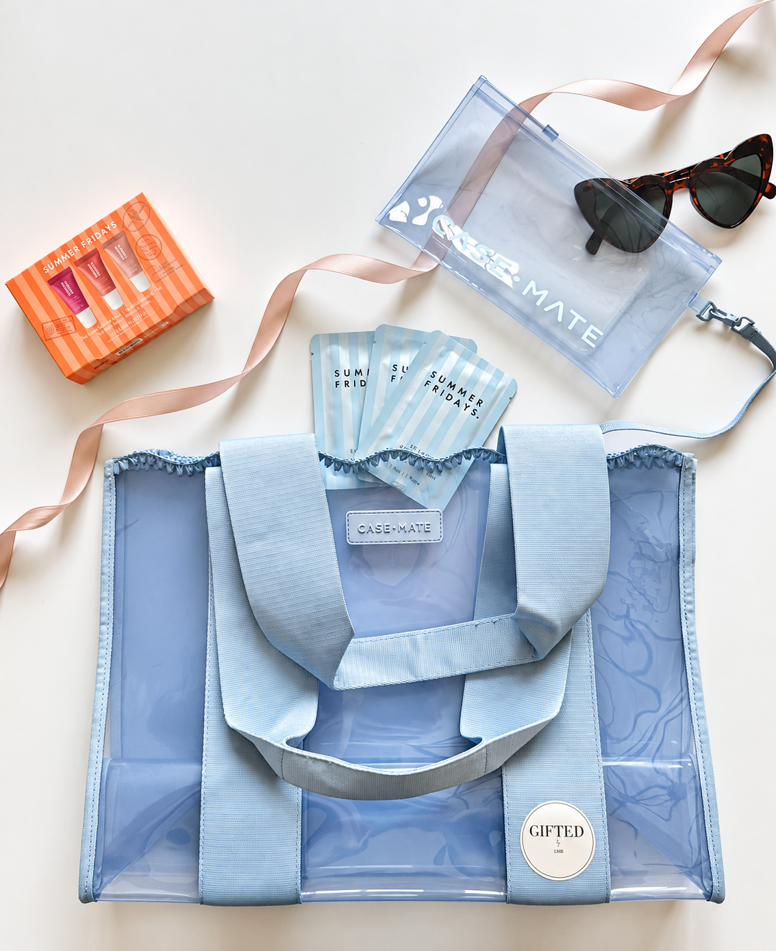 Light blue reusable tote bag with clear panels and handles, containing multiple 'Summer Fridays' skincare sample packets, with a pink ribbon, a clear CASE MATE pouch, and a pair of tortoiseshell sunglasses nearby, all on a white surface.