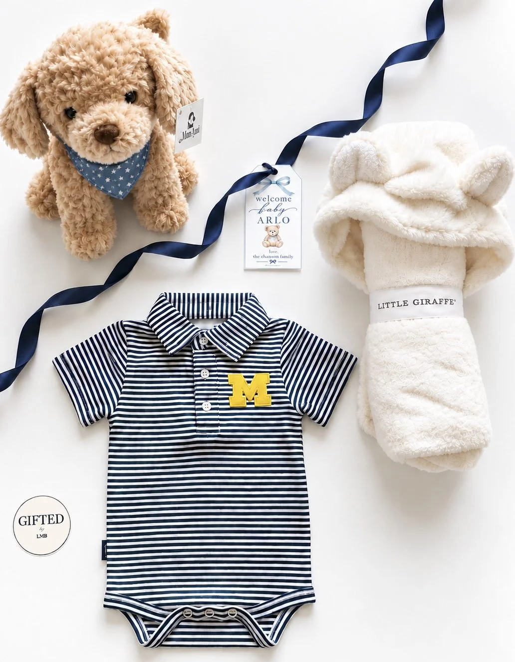 A plush teddy bear with a blue bandana, a striped baby bodysuit with a yellow 'M' patch, a plush white giraffe hooded towel, and a welcome baby tag for Arlo with a teddy bear illustration, arranged on a white surface