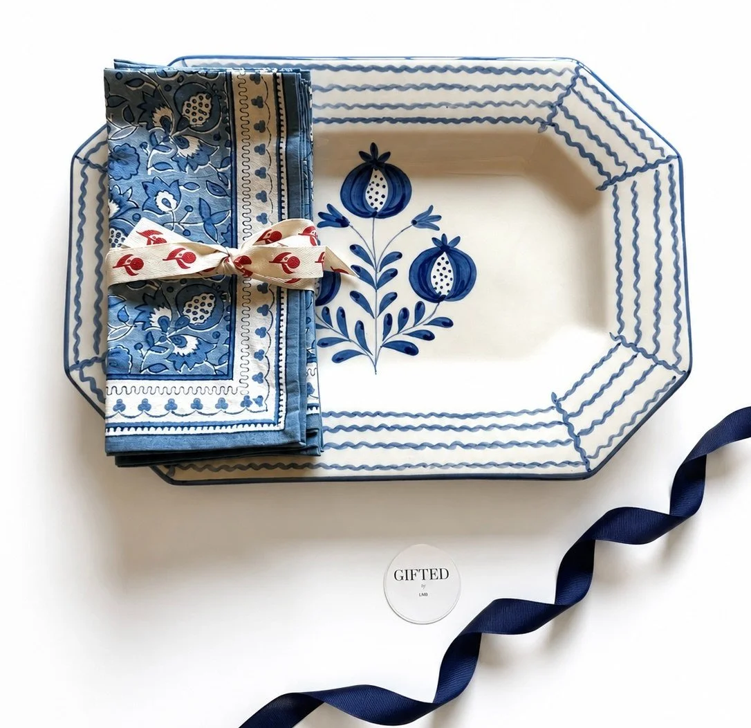 A decorative rectangular platter with blue floral patterns, a blue and white napkin tied with a beige ribbon with red designs, navy blue ribbon, and a small