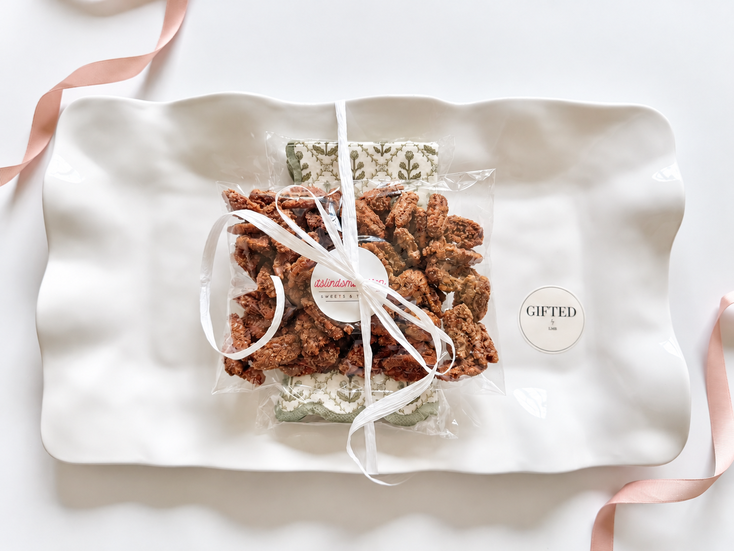 A decorative white ceramic tray holding a clear plastic bag of chocolate or caramel popcorn tied with a white ribbon, with a pillow behind it and a round sticker on the tray that reads "Gifted."