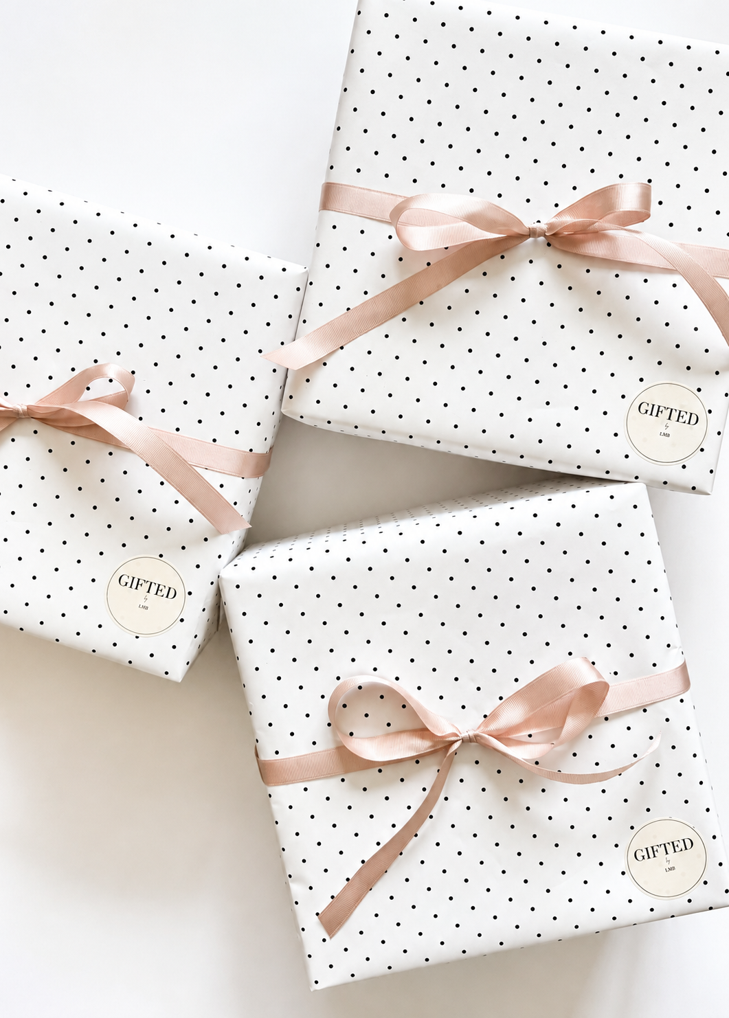 Three gift-wrapped presents with white paper decorated with small black polka dots, tied with pale pink satin ribbons, on a white surface.