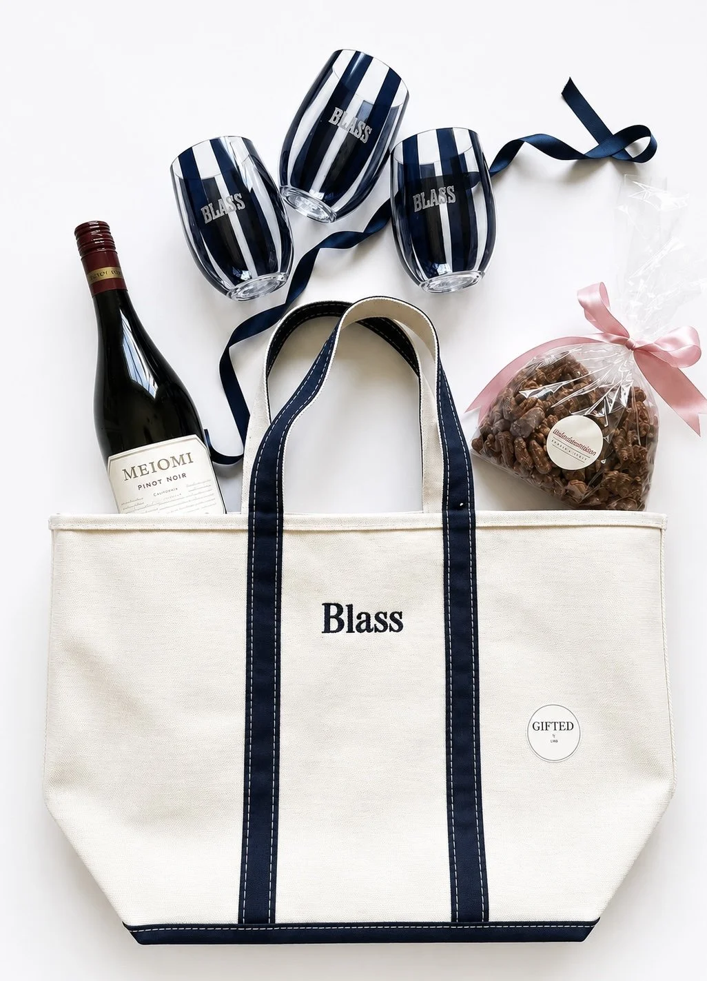 A white tote bag with navy blue handles and trim, with the word 'Blass' on it, surrounded by a bottle of wine, three navy blue striped glasses labeled 'Blass,' a bag of chocolates with a pink bow, and a navy blue ribbon.
