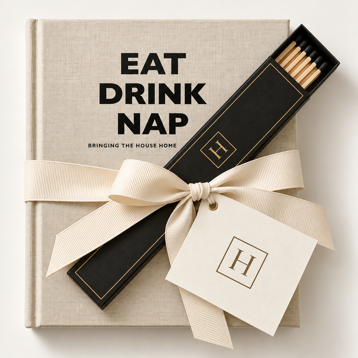 A gift set with a book titled 'Eat Drink Nap', a box of matches by H, and a cream-colored ribbon with a gift tag, all arranged on a beige textured surface.