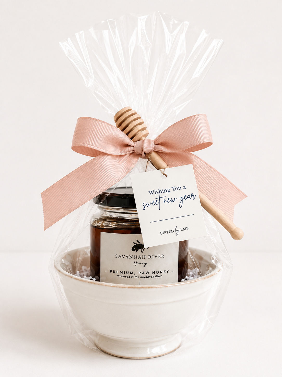 A gift set of raw honey in a jar with a label, wrapped in clear plastic with a pink ribbon, a wooden honey dipper, and a tag that says 'Wishing You a sweet new year' on a white background.