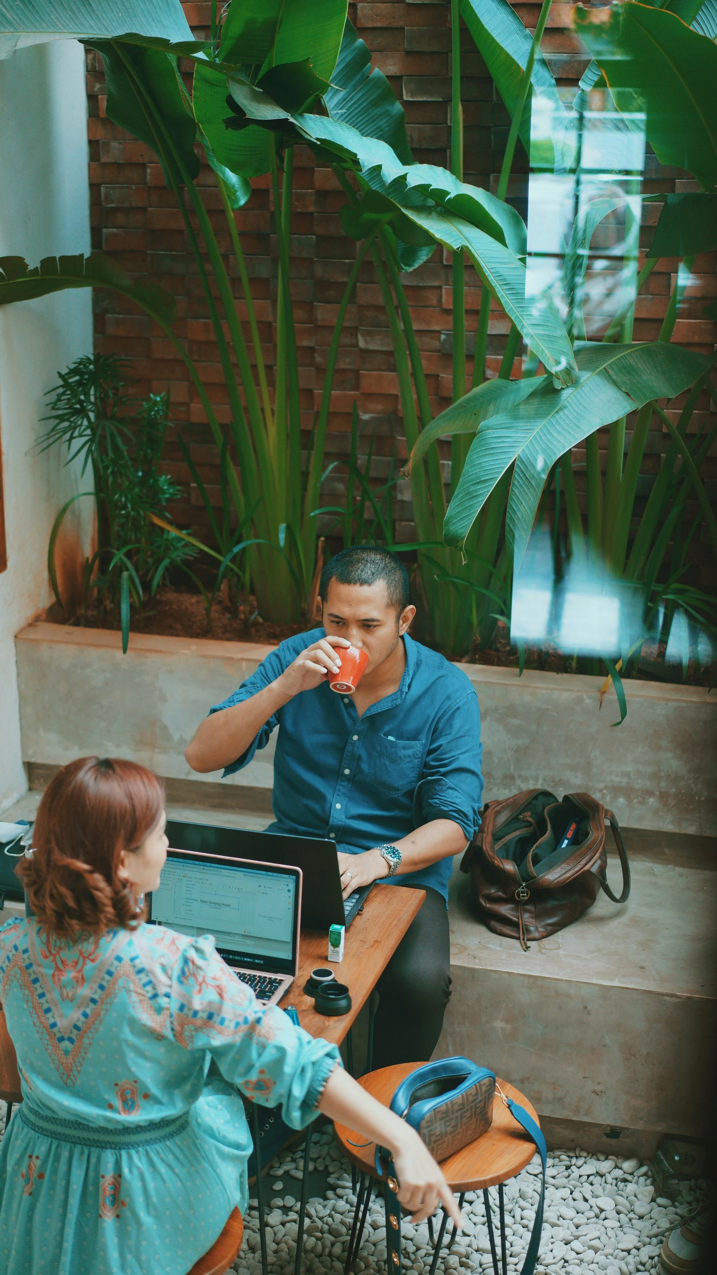 A man sitting at a table drinking coffee or tea, with a woman sitting across from him working on a laptop, in an indoor plant-filled setting.