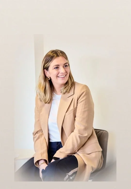 A woman with shoulder-length blonde hair, wearing a beige blazer over a white top, sitting on a chair and smiling.