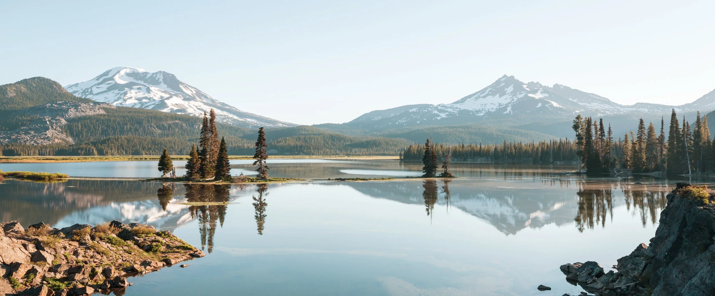 Manufactured Home Communities in Oregon amongst Snow-capped mountains over a calm lake surrounded by pine trees and rocky shores.