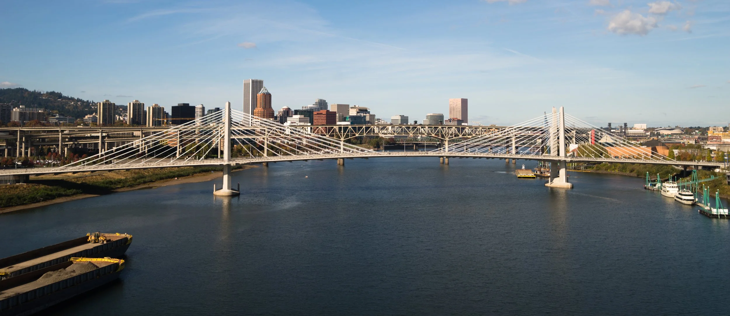 Portland, Oregon, Manufactured Home Communities Advisory Groups. Portland city skyline with a cable-stayed bridge over a river, boats docked on the right, and a clear sky.