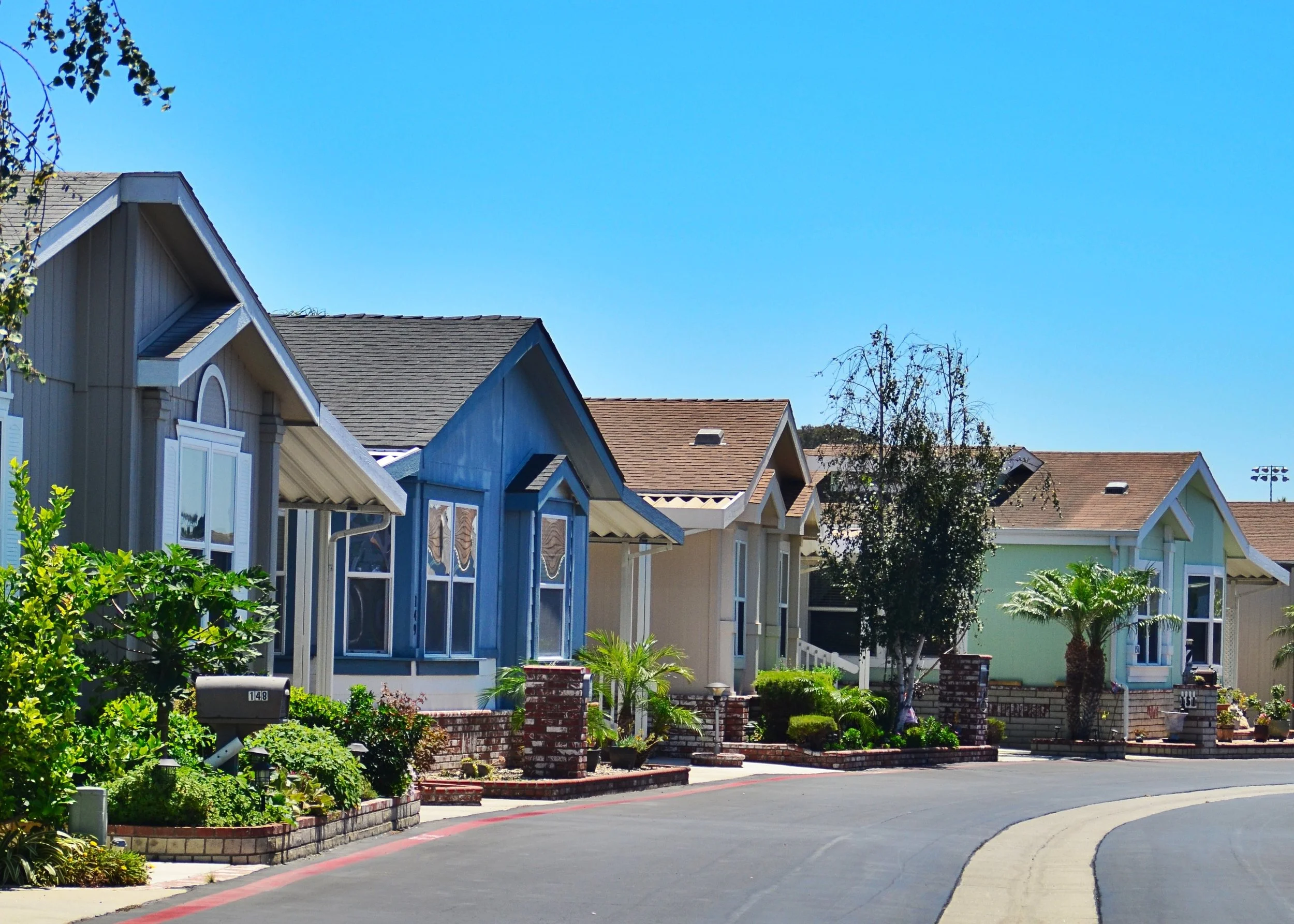 Line of colorful houses with well-maintained gardens in front, under a clear blue sky. The houses have different shades of blue, green, and beige, and feature gabled roofs with shingles. New Manufactured Homes, Mobile Home Park, Oregon