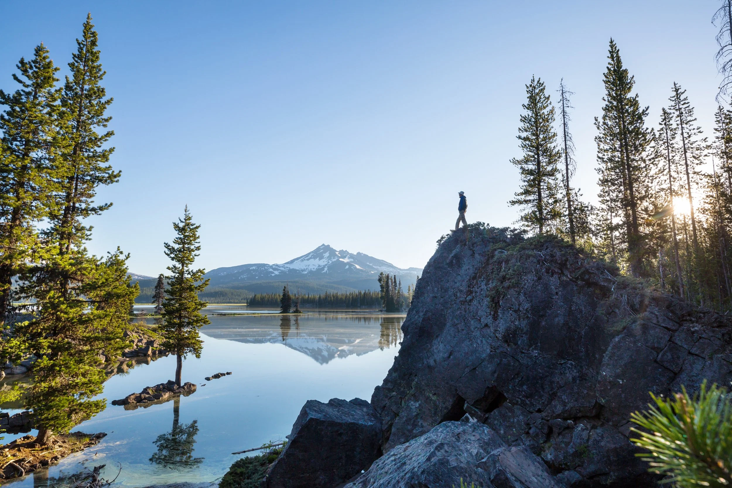 A person standing on a rocky outcrop surrounded by tall evergreen trees with a mountain and lake in the background during sunset. Save time, enjoy vacations, allow someone to help you make your mobile home community profitable
