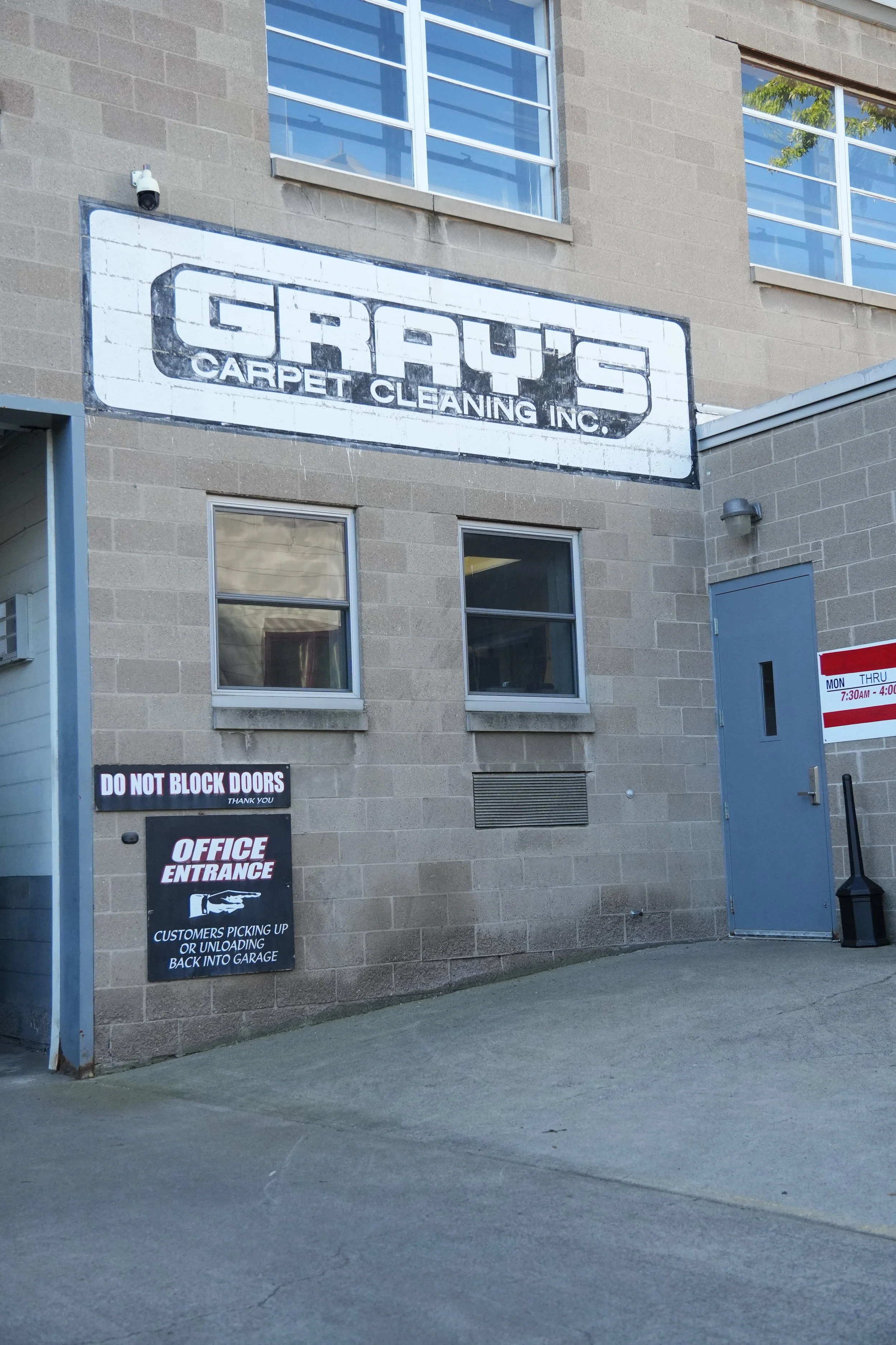 The exterior of a carpet cleaning business named 'GRAYS Carpet Cleaning Inc.' shows a large sign on a brick wall, with smaller signs below indicating the office entrance and instructions for waiting customers. There are two windows above the sign and a blue door to the right.