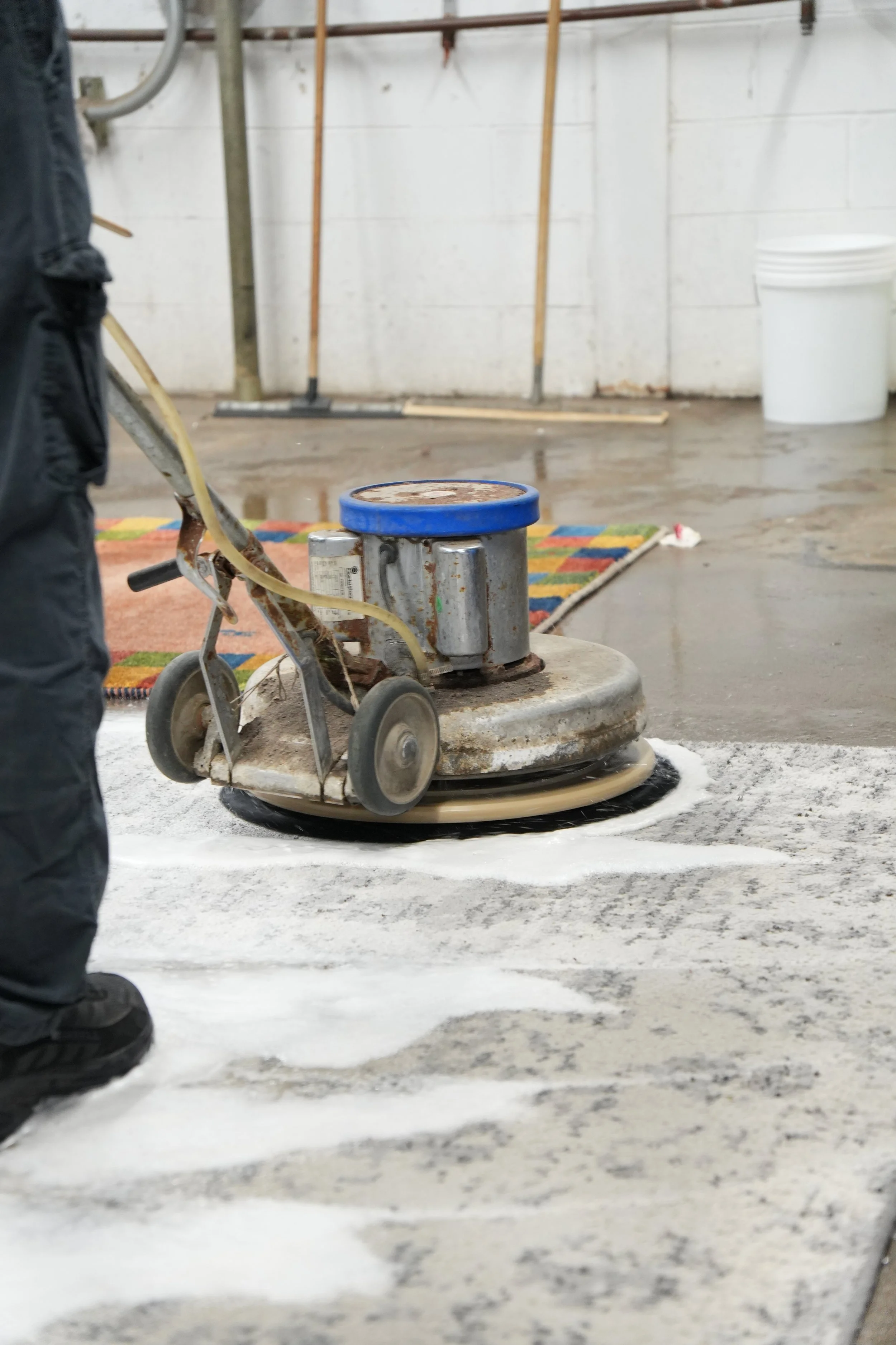Rug Cleaning Machine machine operating on concrete floor, with person standing nearby and colorful rug in background.