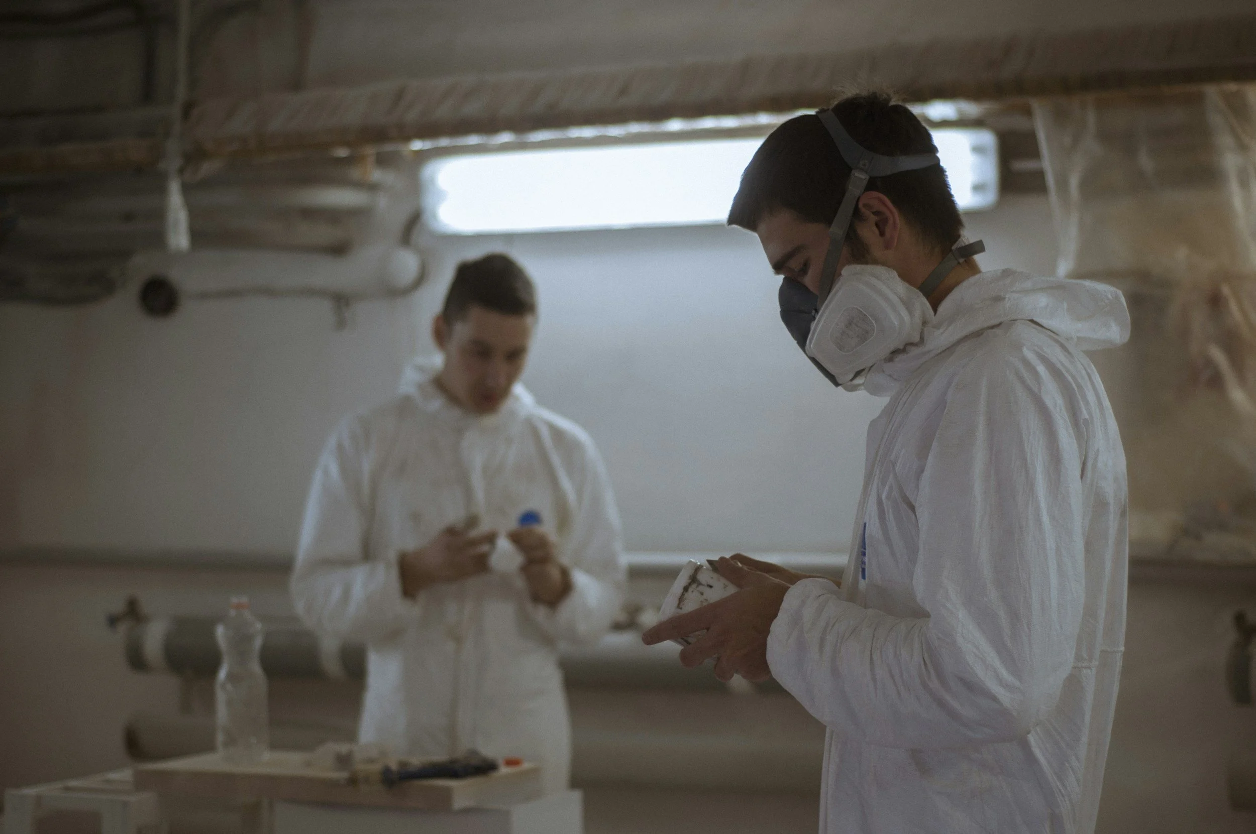 Two individuals wearing protective masks and white coveralls in a room with scientific equipment and supplies, possibly a laboratory or research facility.