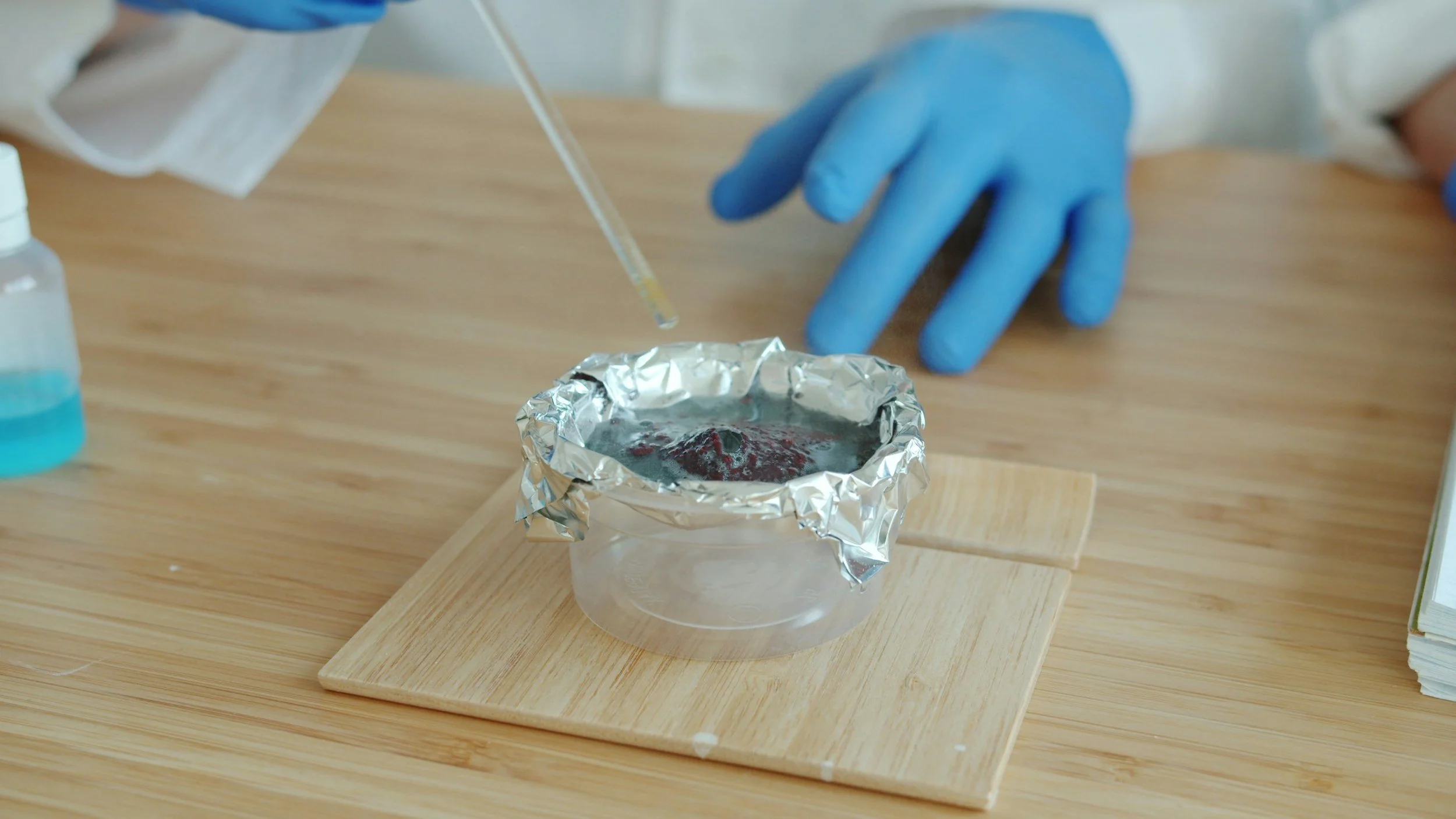 Person wearing blue gloves handling a small container covered with aluminum foil, with a dropper or pipette nearby, on a wooden surface.