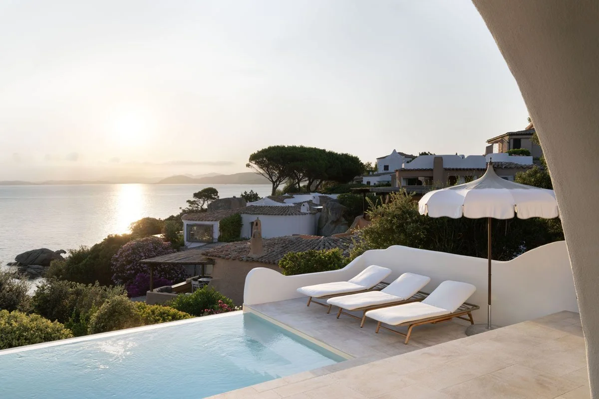 Luxury coastal villa with swimming pool, lounge chairs, and umbrella overlooking a sunset by the water with trees and homes in the background.