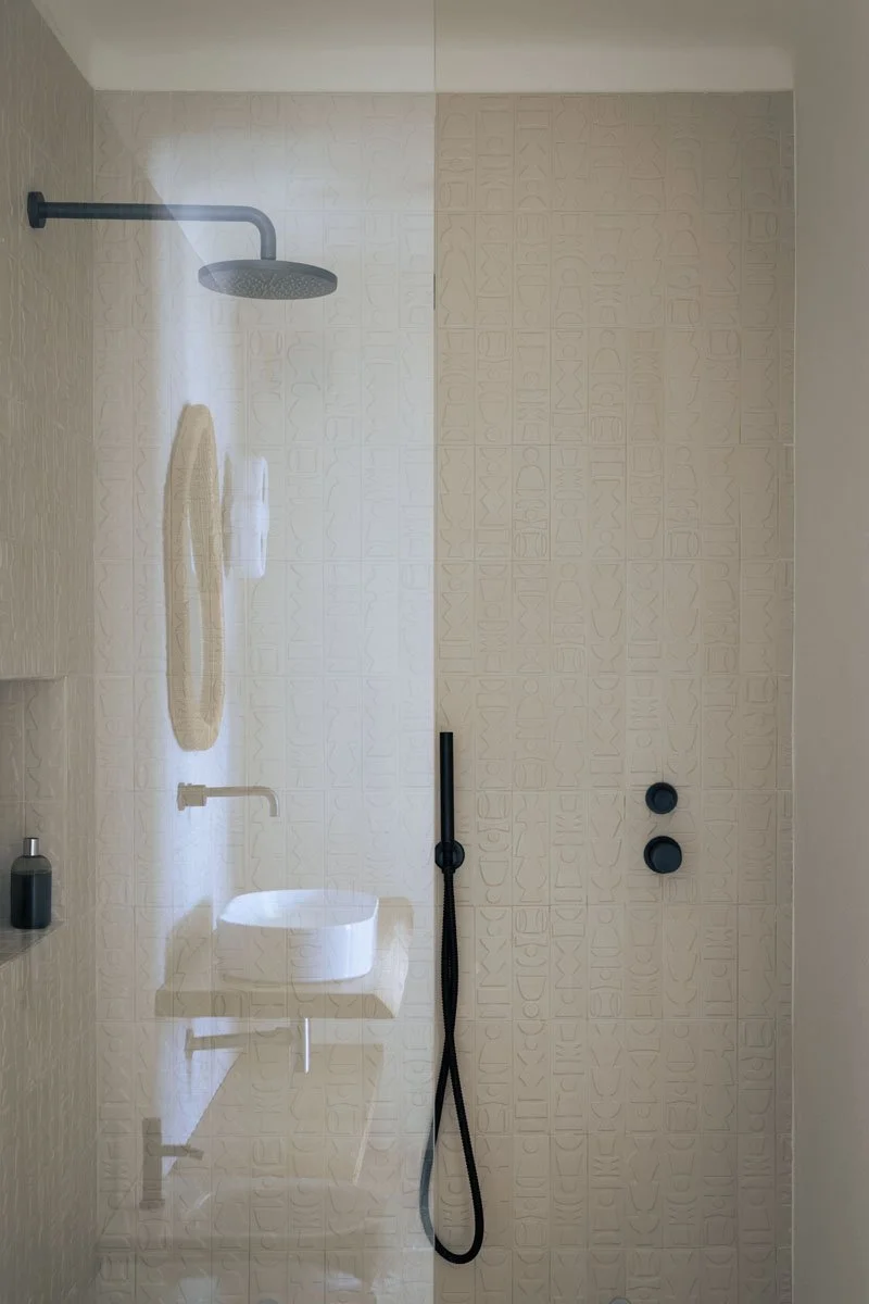 Modern walk-in shower with a rain showerhead, a built-in soap dispenser, a woven bath mat, a small white curved sink, a wall-mounted faucet, and a black handheld shower wand, with textured tiles on the walls.