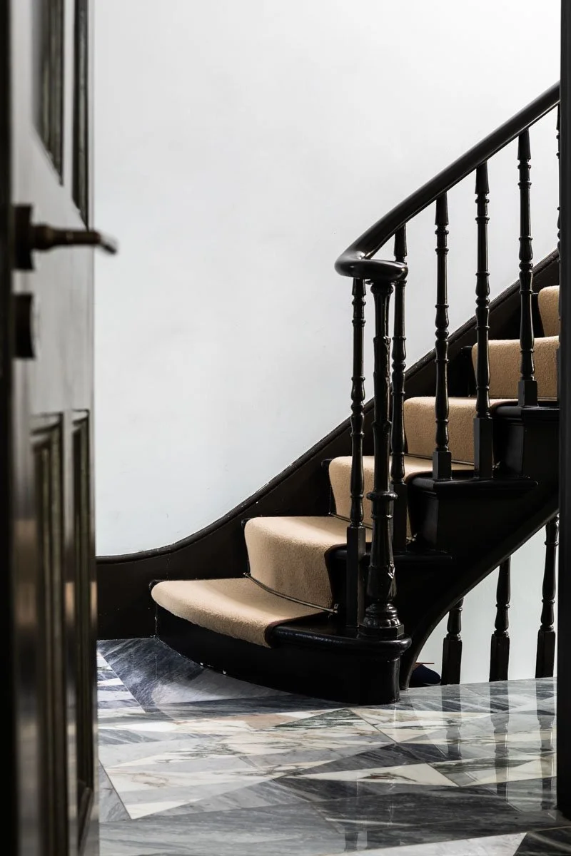 A staircase with black wooden handrail and balusters, beige carpeted steps, and a marble floor at the bottom, with part of a wall visible in the background.