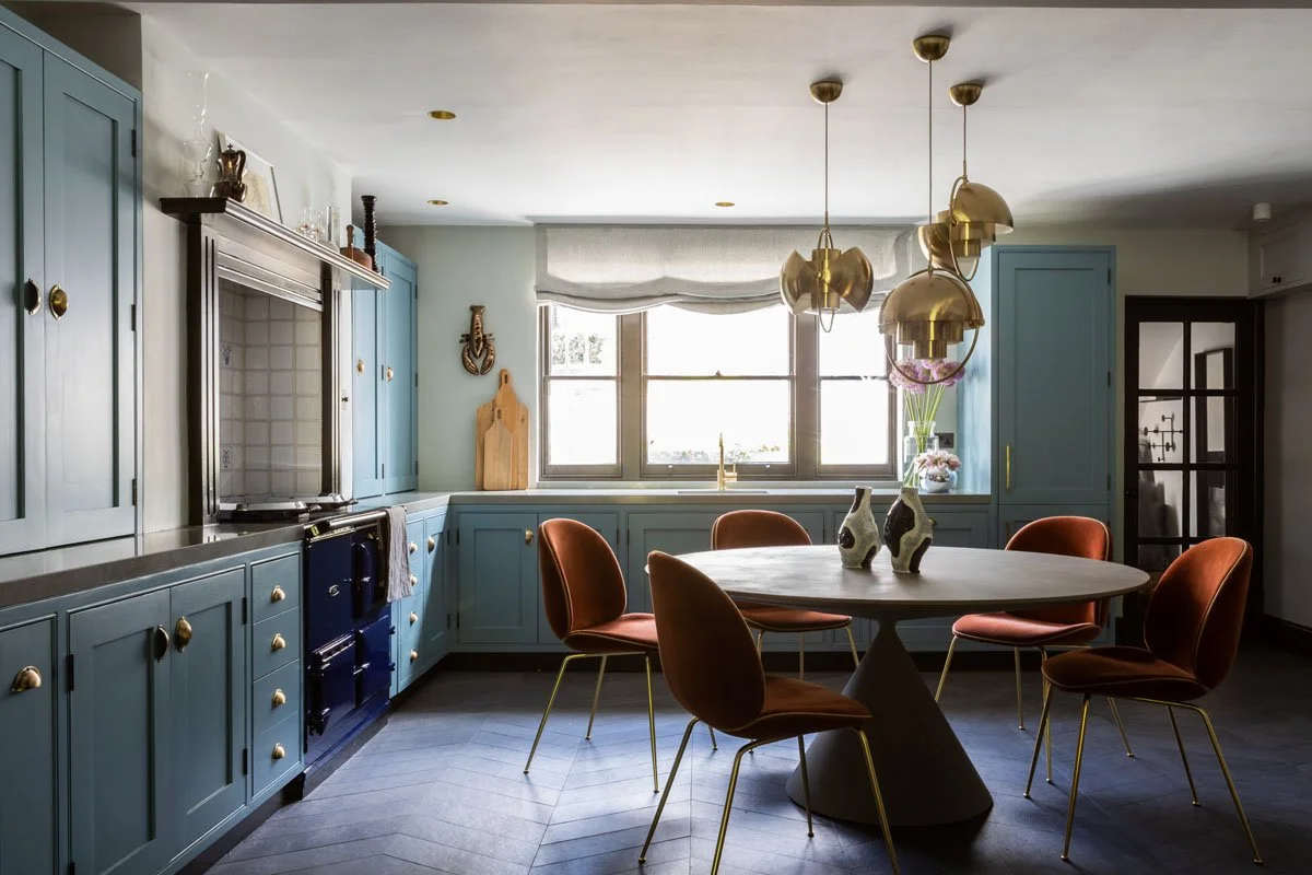 A bright kitchen with blue cabinets, a round dining table with five orange chairs, and gold pendant lights hanging from the ceiling. There are decorative vases and a window with blinds letting in natural light.